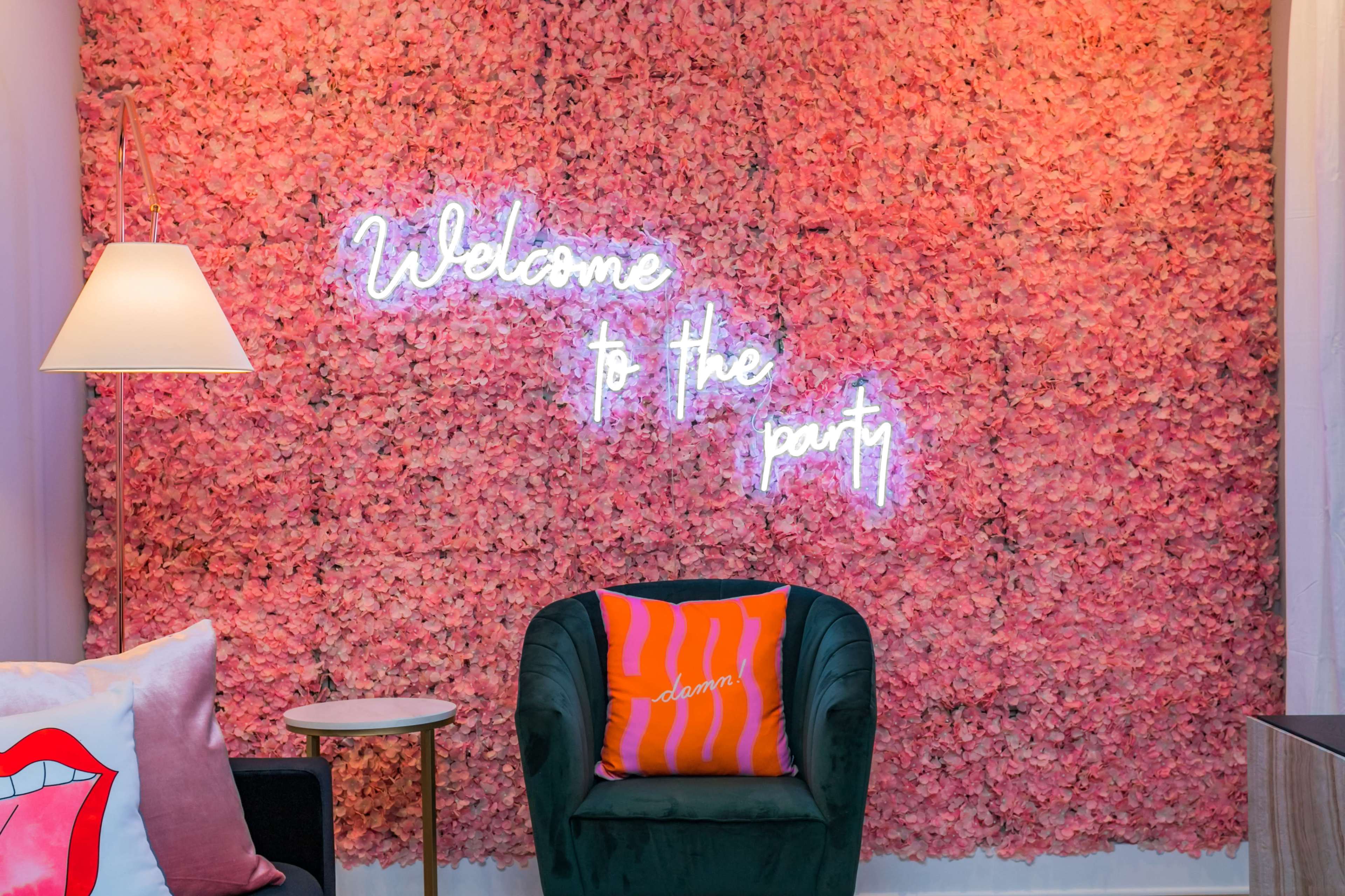 A seating area adorned with a pink floral wall and a neon sign that reads "Welcome to the party."