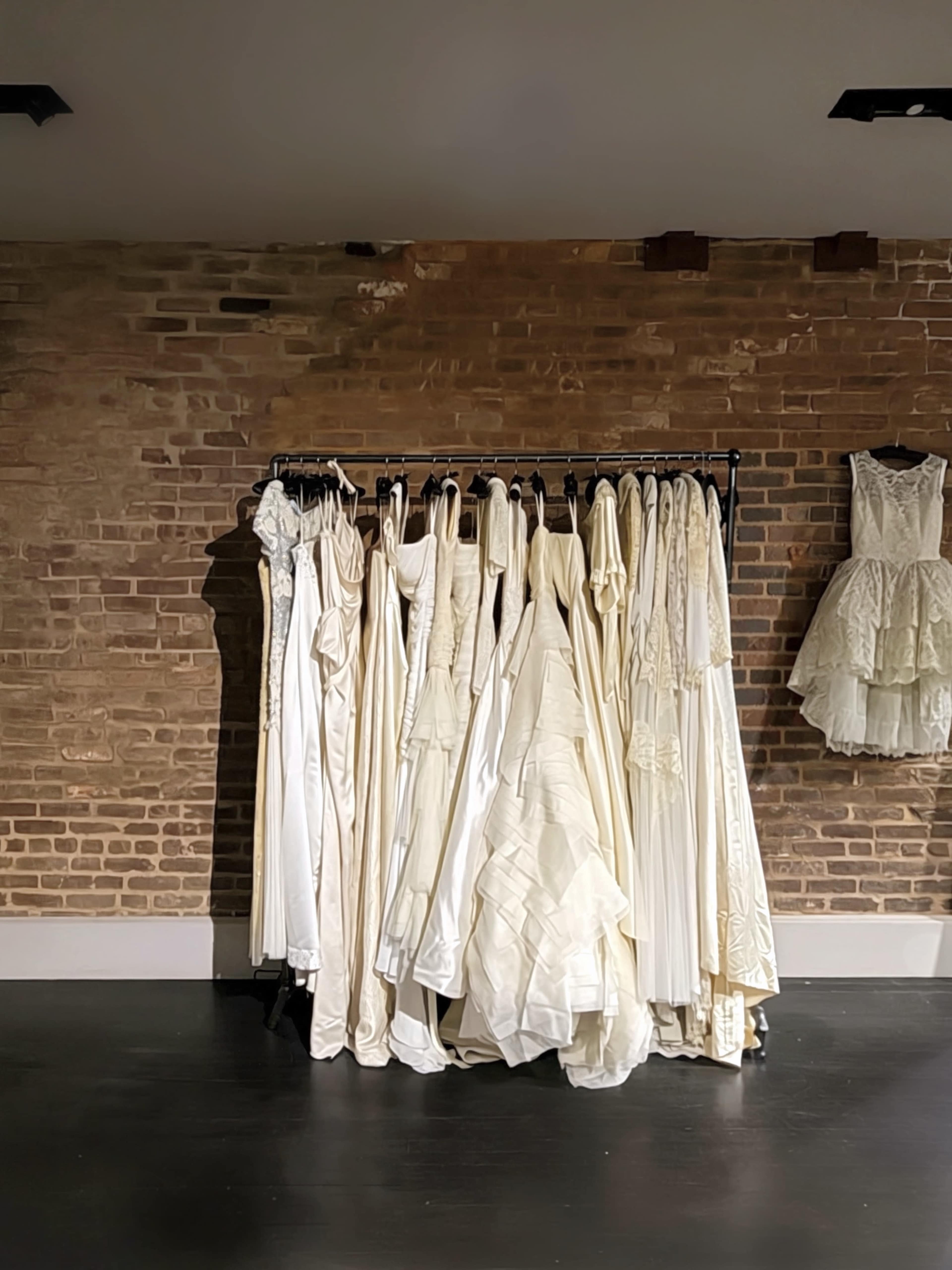A rack displays a variety of white wedding dresses against a brick wall.
