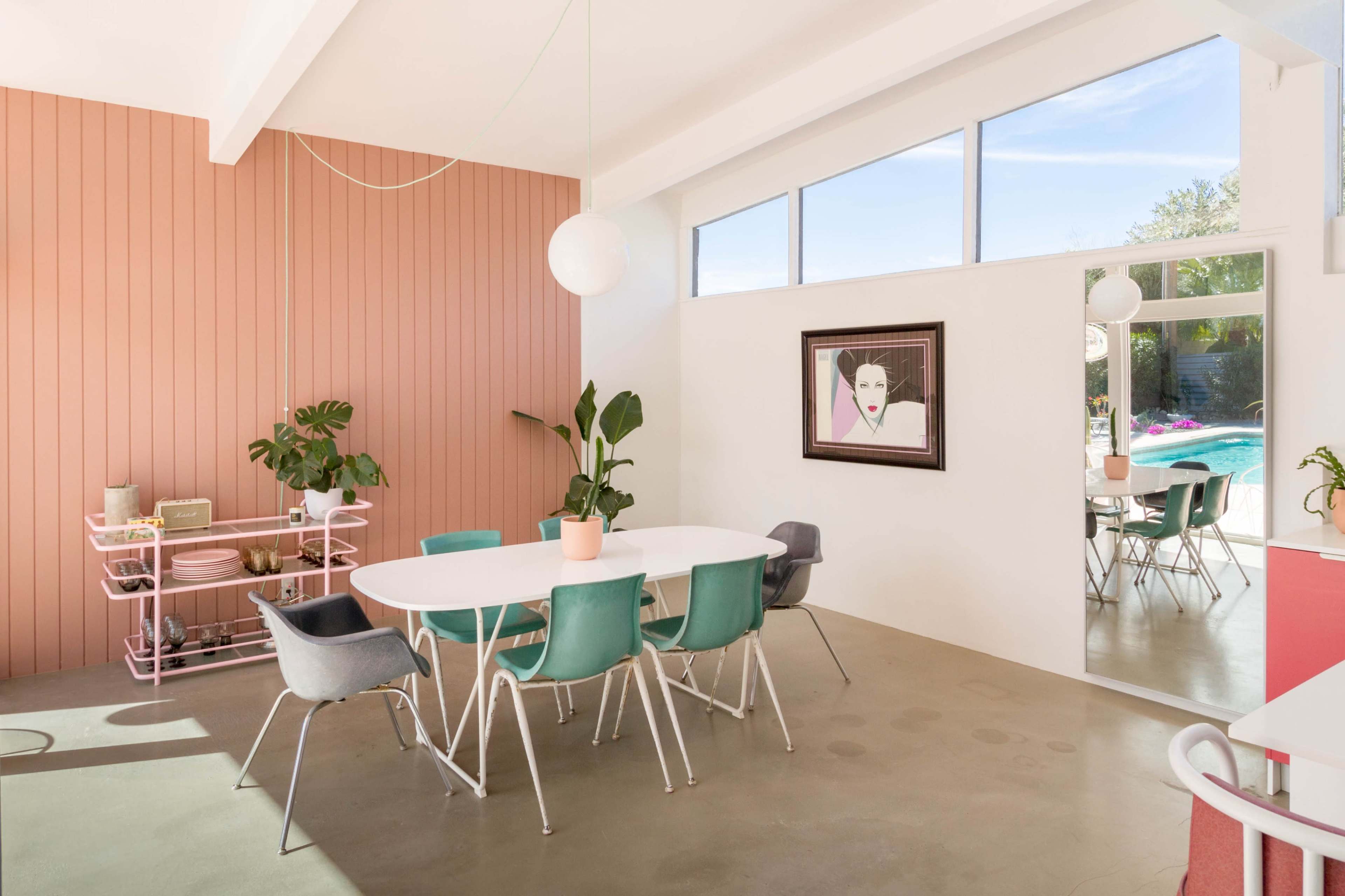 A modern dining area features a round white table surrounded by colorful chairs, with a pink wall, a large window, and a mirror reflecting a poolside view.