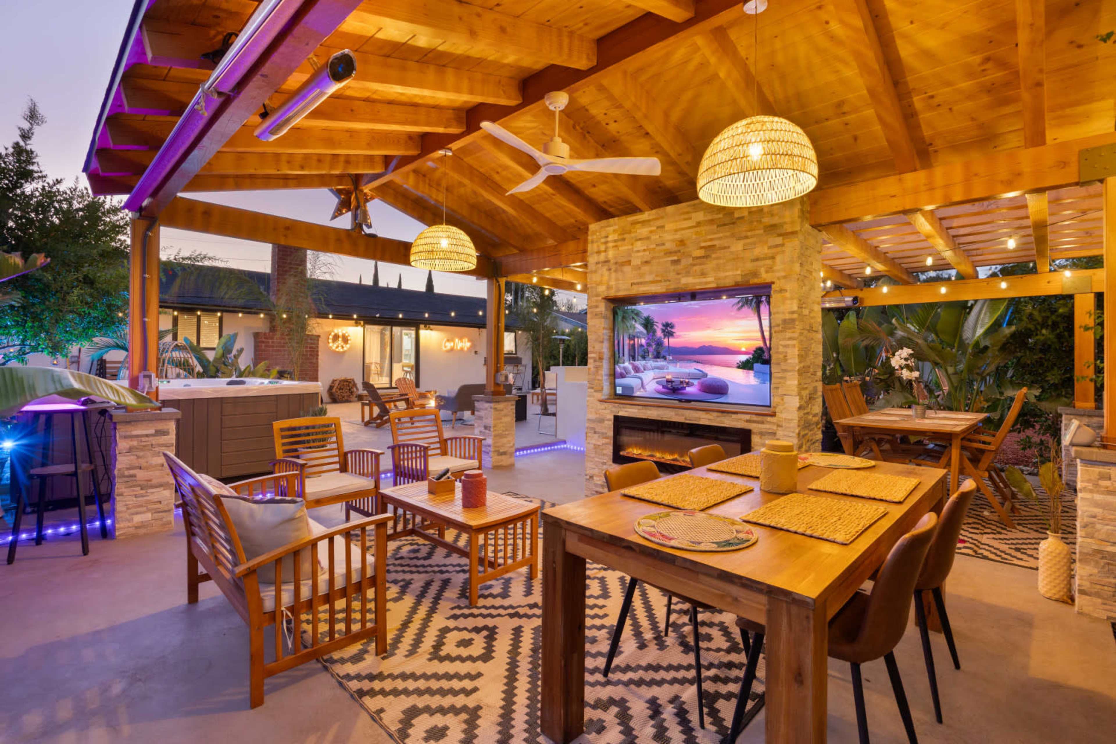 The image shows a cozy outdoor seating area with wooden furniture, string lights, and a large screen displaying a scenic view.