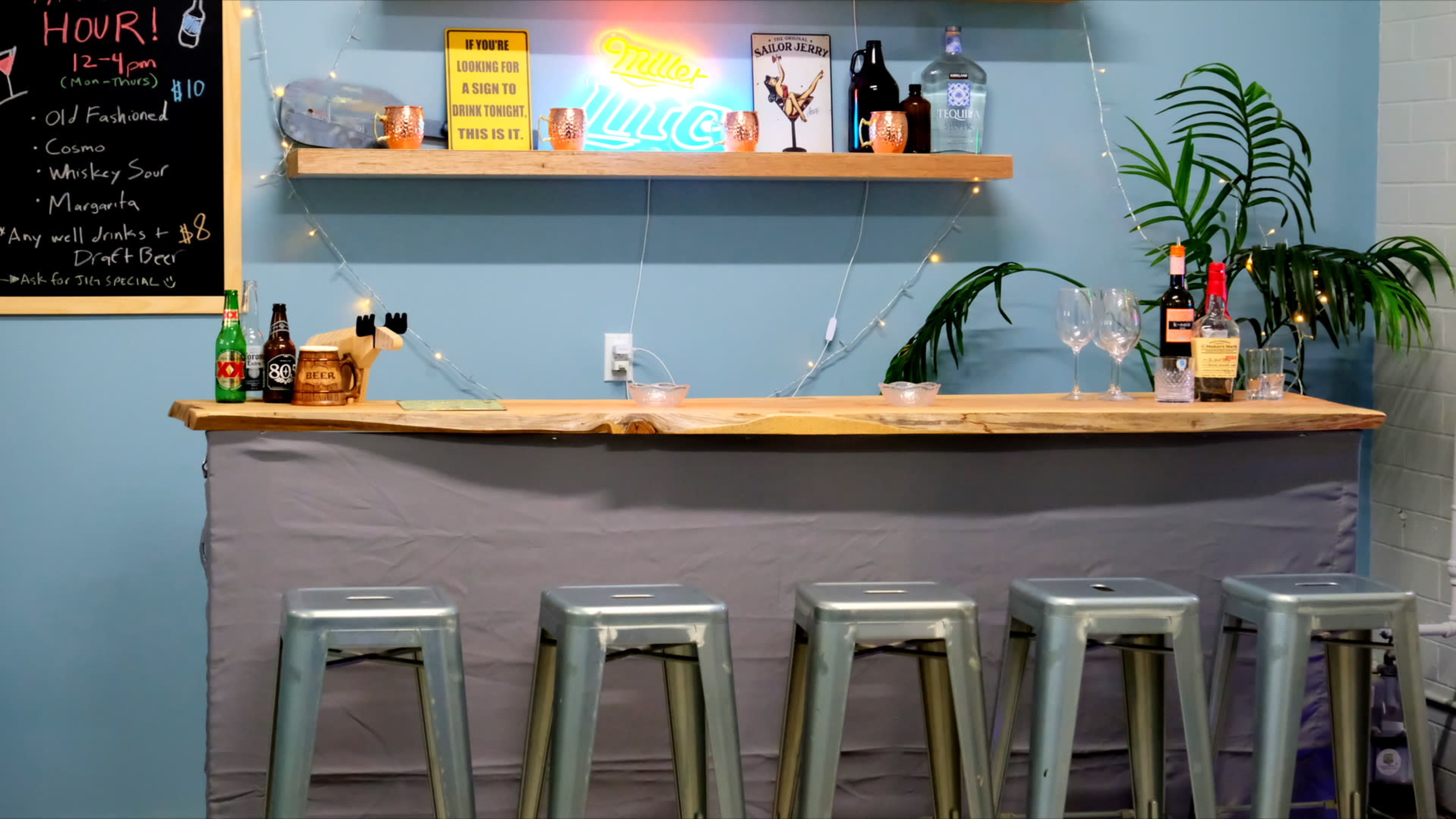 A bar area features a wooden countertop with several bottles of liquor, glasses, and decorative items, accompanied by metallic stools lined up in front.