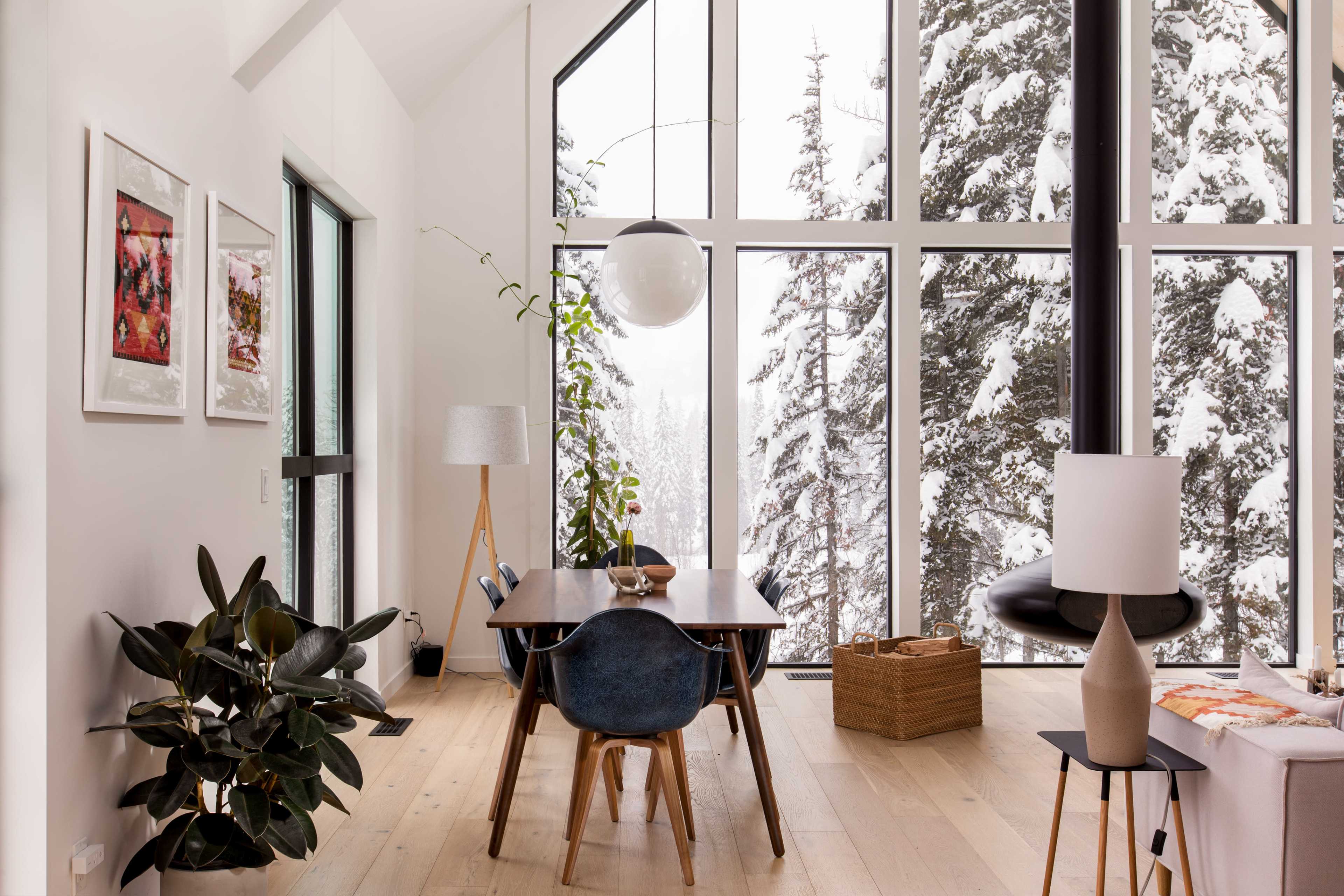 A modern dining area features a wooden table surrounded by chairs, with large windows displaying a snowy landscape outside.