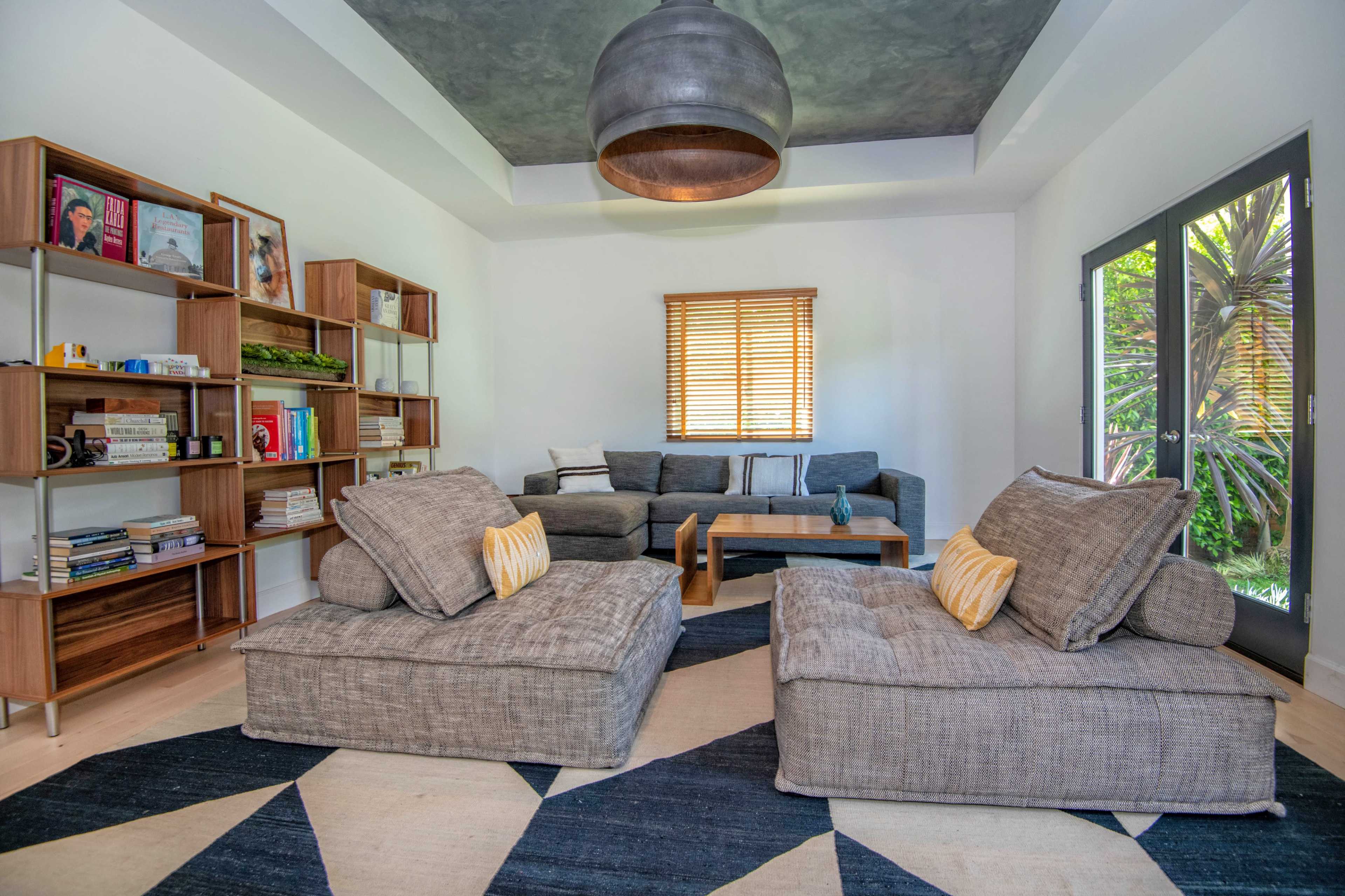 A modern living room featuring two large, gray lounge chairs, a wooden coffee table, bookshelves filled with books, and a large window with blinds.