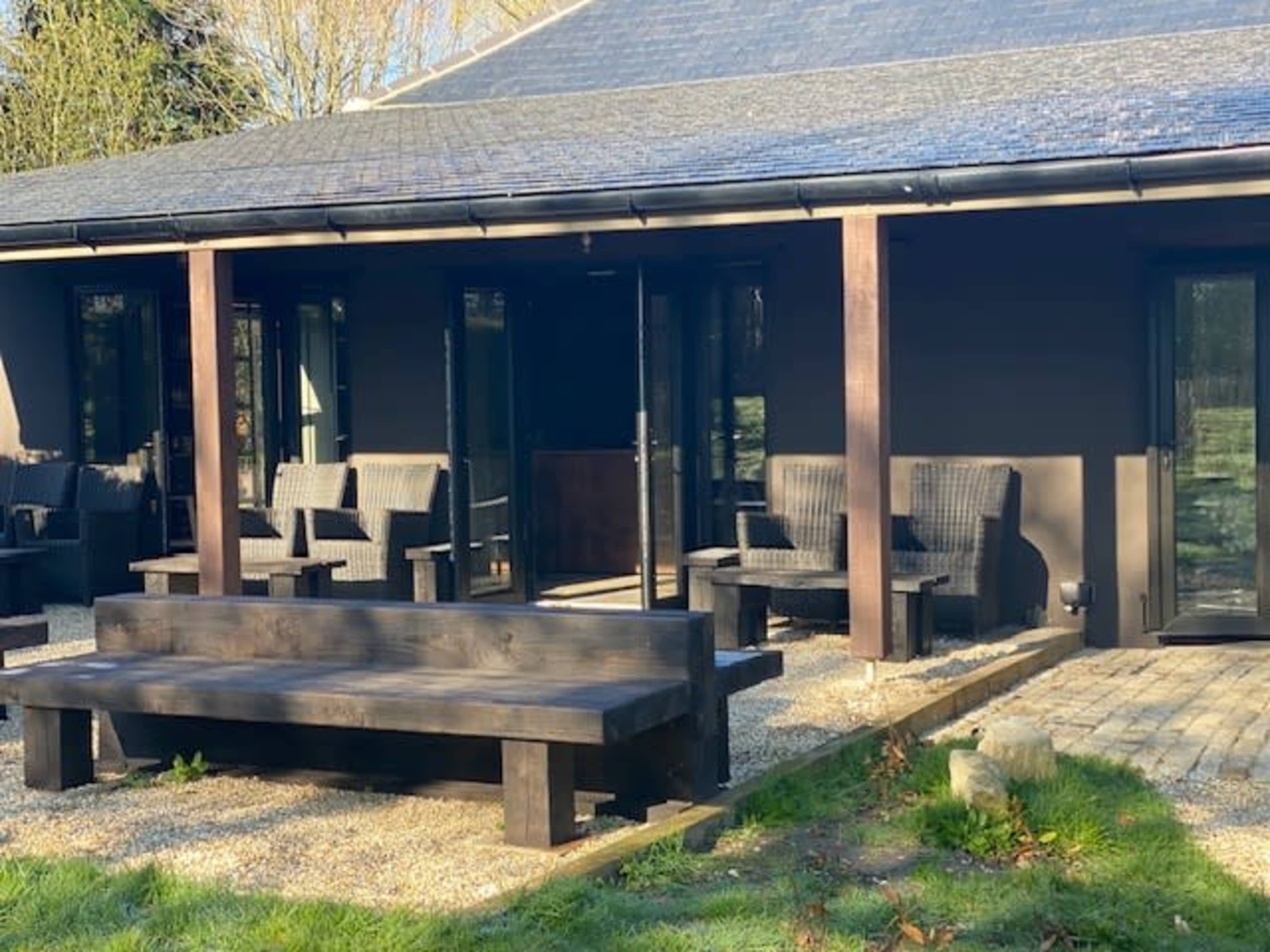 The image shows a modern building with a dark exterior and a front patio featuring wooden benches and wicker chairs.