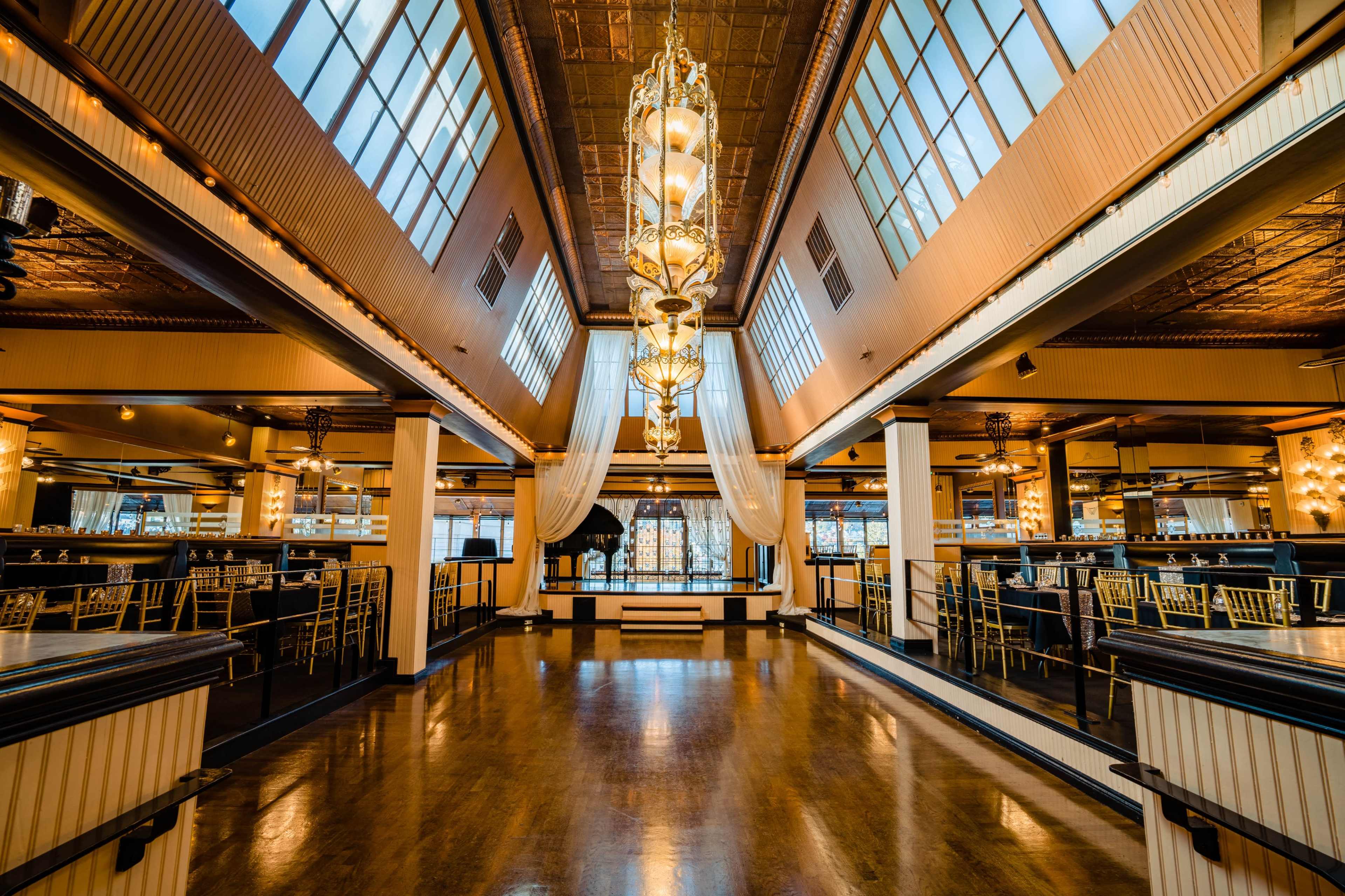The image shows a spacious, elegantly decorated restaurant interior with large windows, chandeliers, and a polished wooden floor.