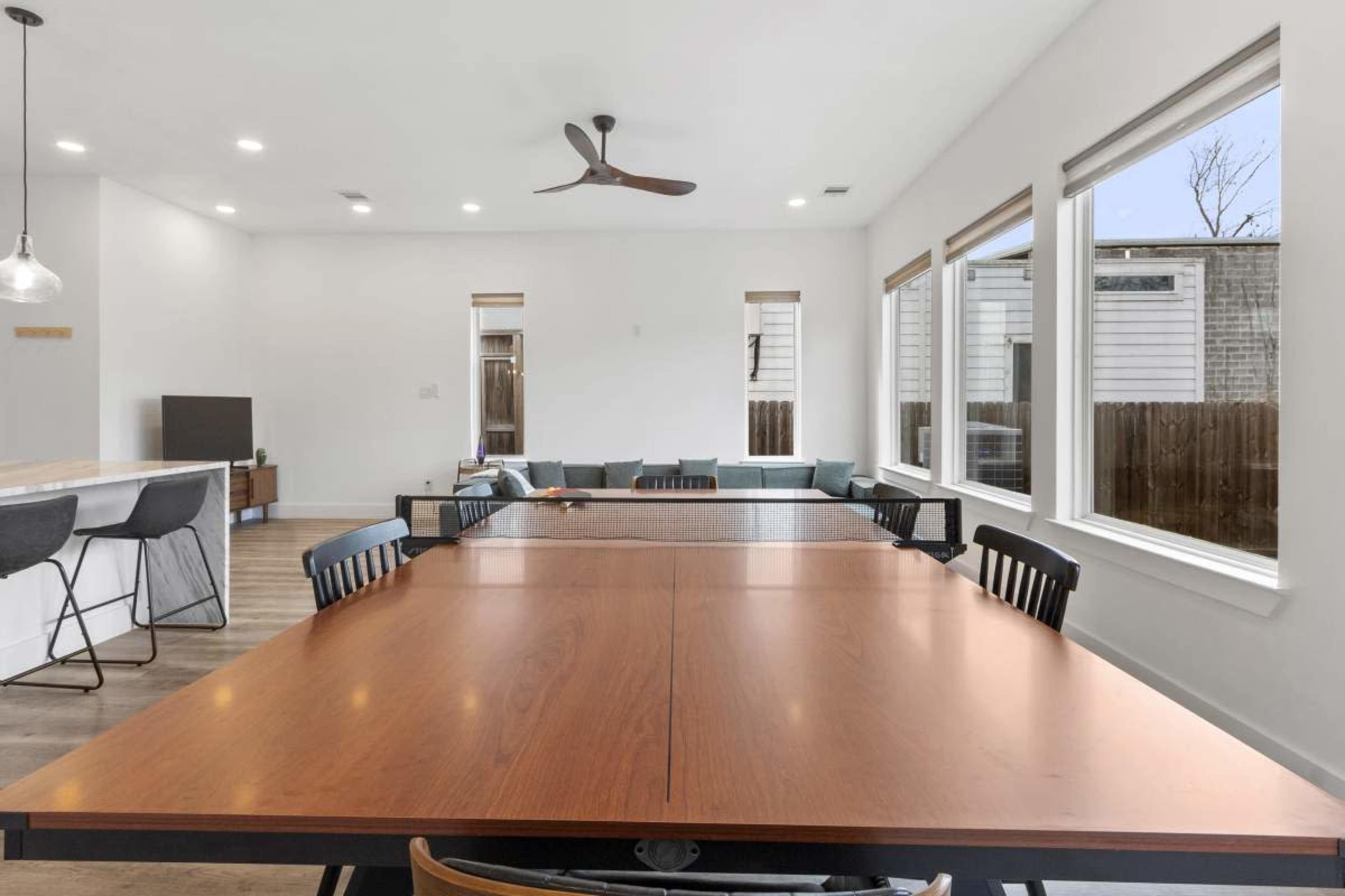 The image shows a modern dining area with a large wooden table surrounded by black chairs, leading to a spacious living room with large windows.