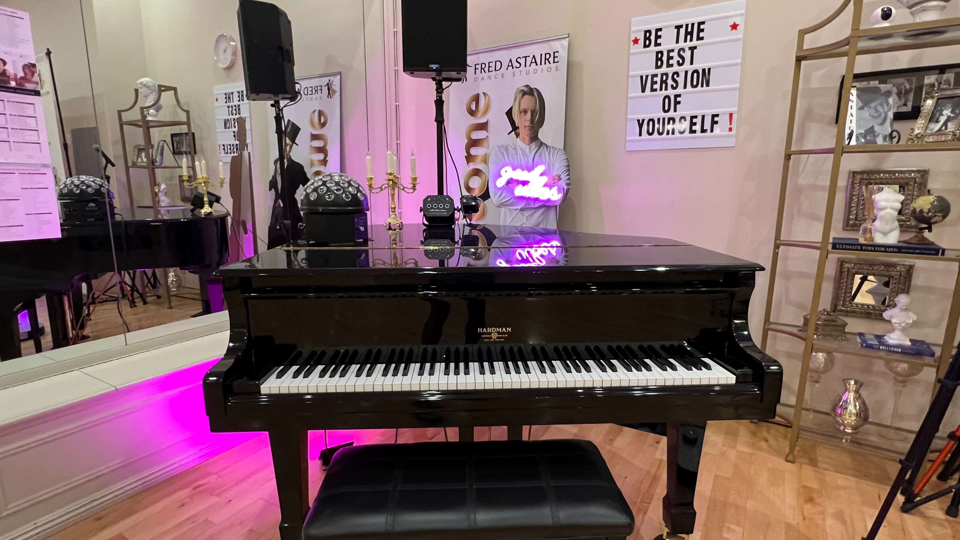 A black grand piano in a studio setting, accompanied by mirrors, decorative shelves, and a motivational sign on the wall.