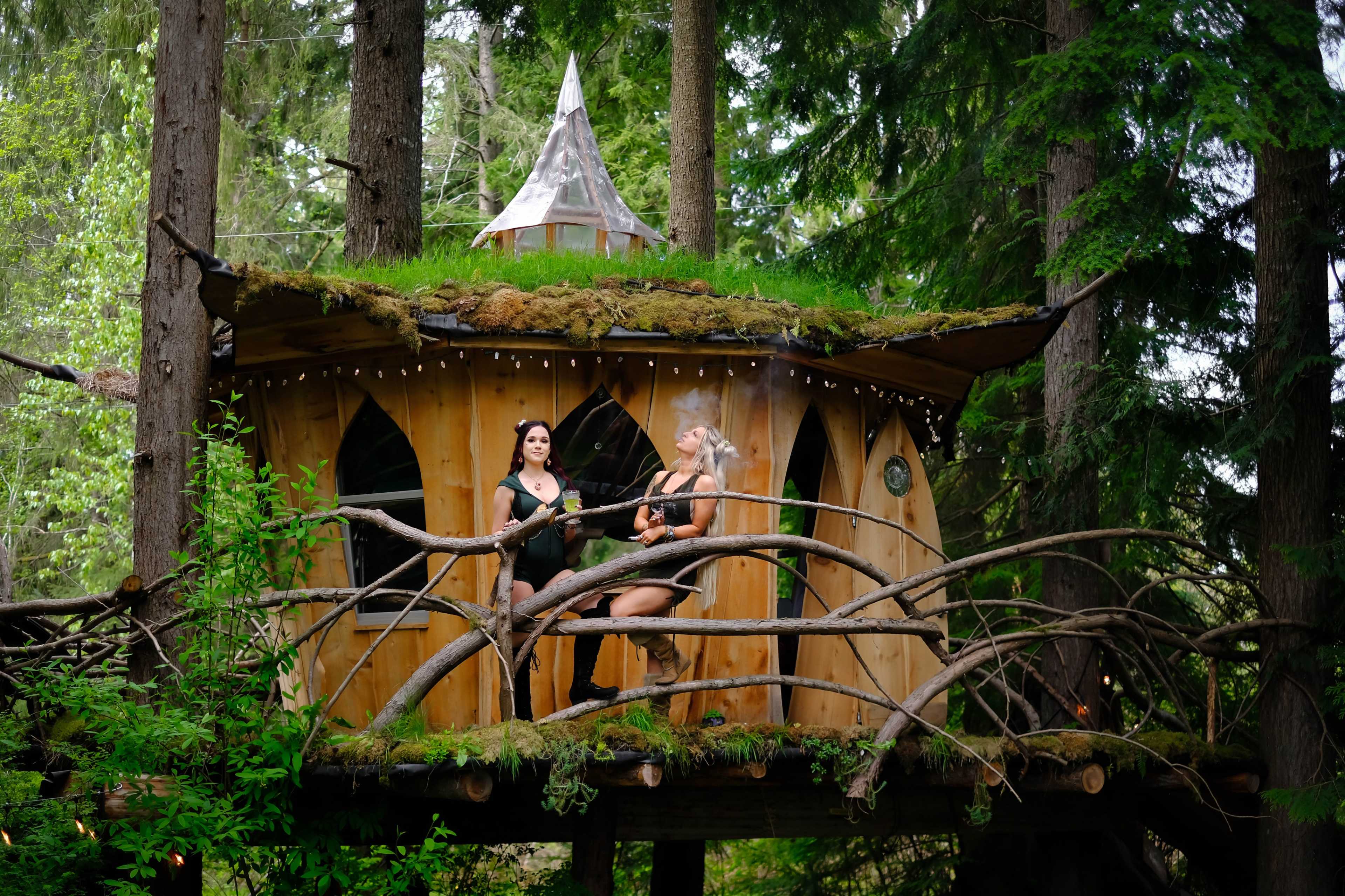 Two women are playing instruments on the balcony of a wooden treehouse surrounded by dense greenery.