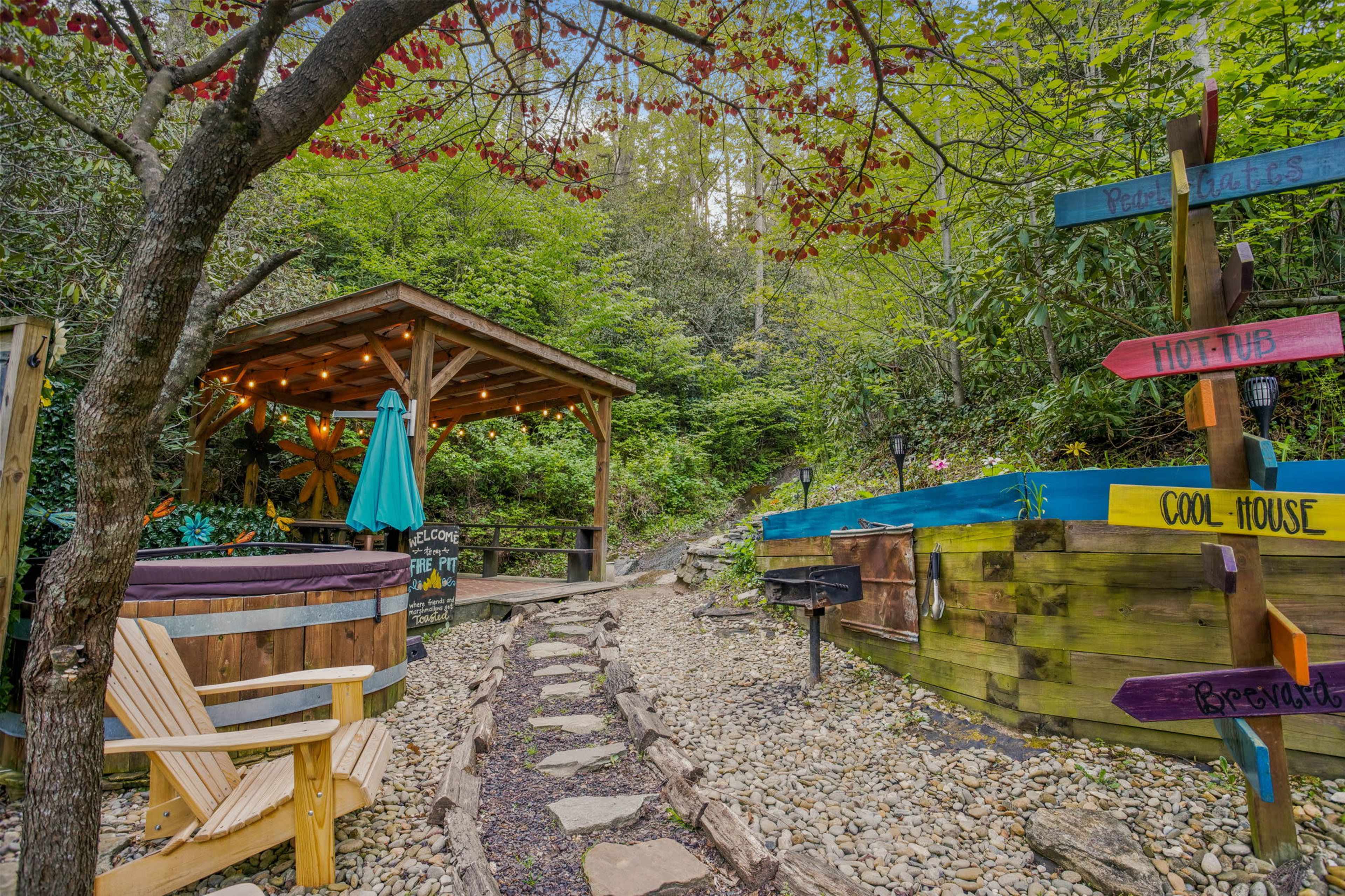 A wooden gazebo with string lights sits near a path leading to a colorful signpost and a small area featuring a hot tub.