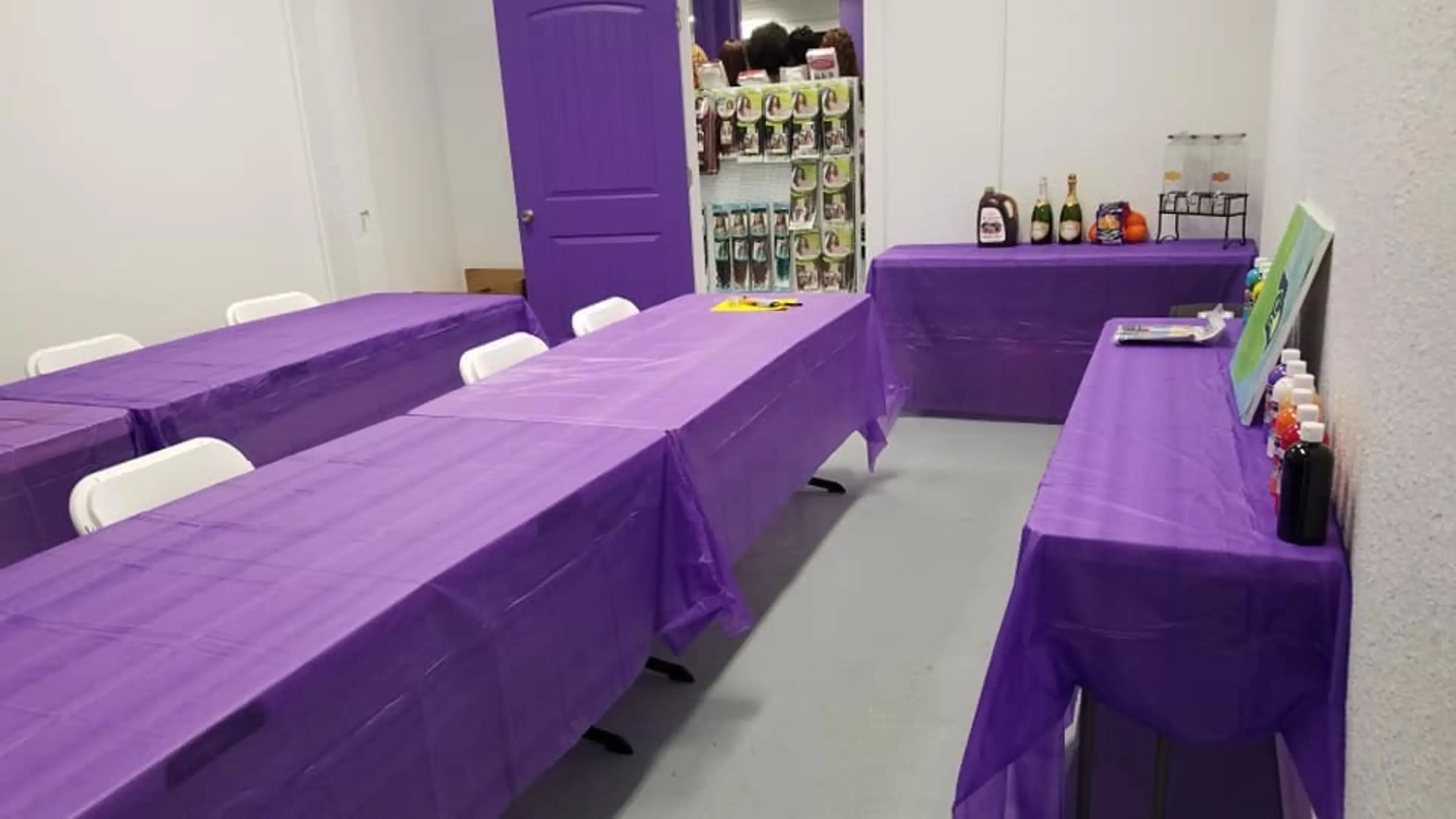The image shows a room with several tables covered in purple tablecloths, arranged in a setup suitable for an event, with a wall displaying colorful items in the background.