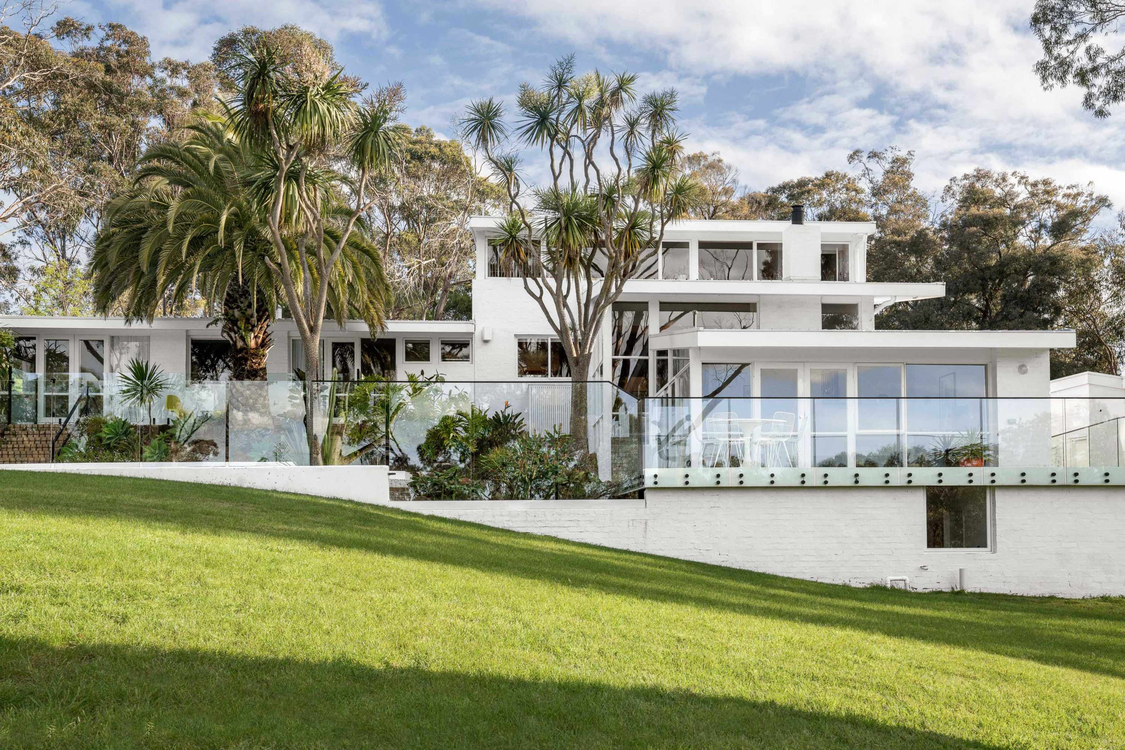 The image features a modern white house with large glass windows, situated on a grassy hillside surrounded by trees and tropical plants.