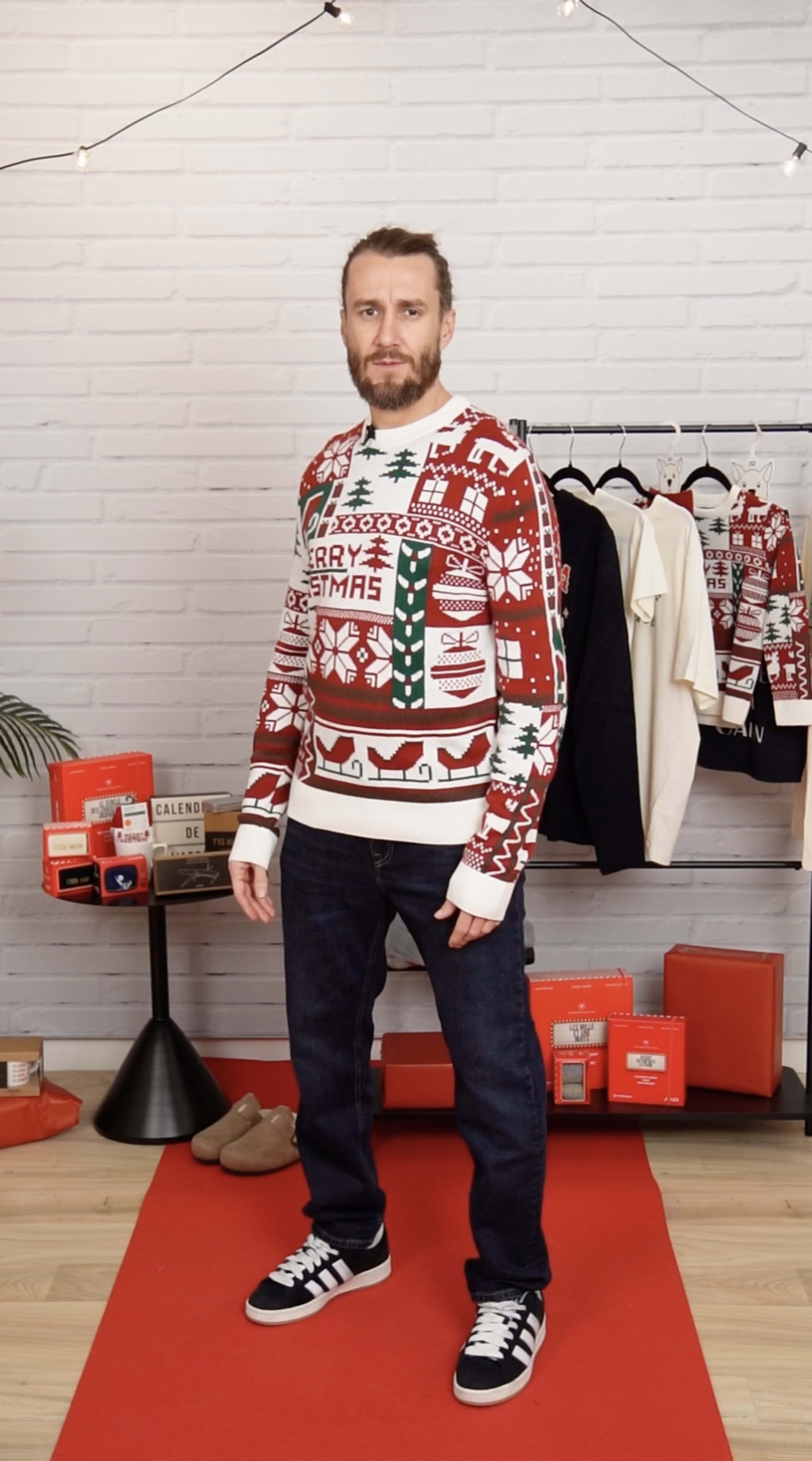 A man stands in a festive setting wearing a colorful Christmas sweater, paired with dark jeans and sneakers, surrounded by seasonal decorations and clothing.