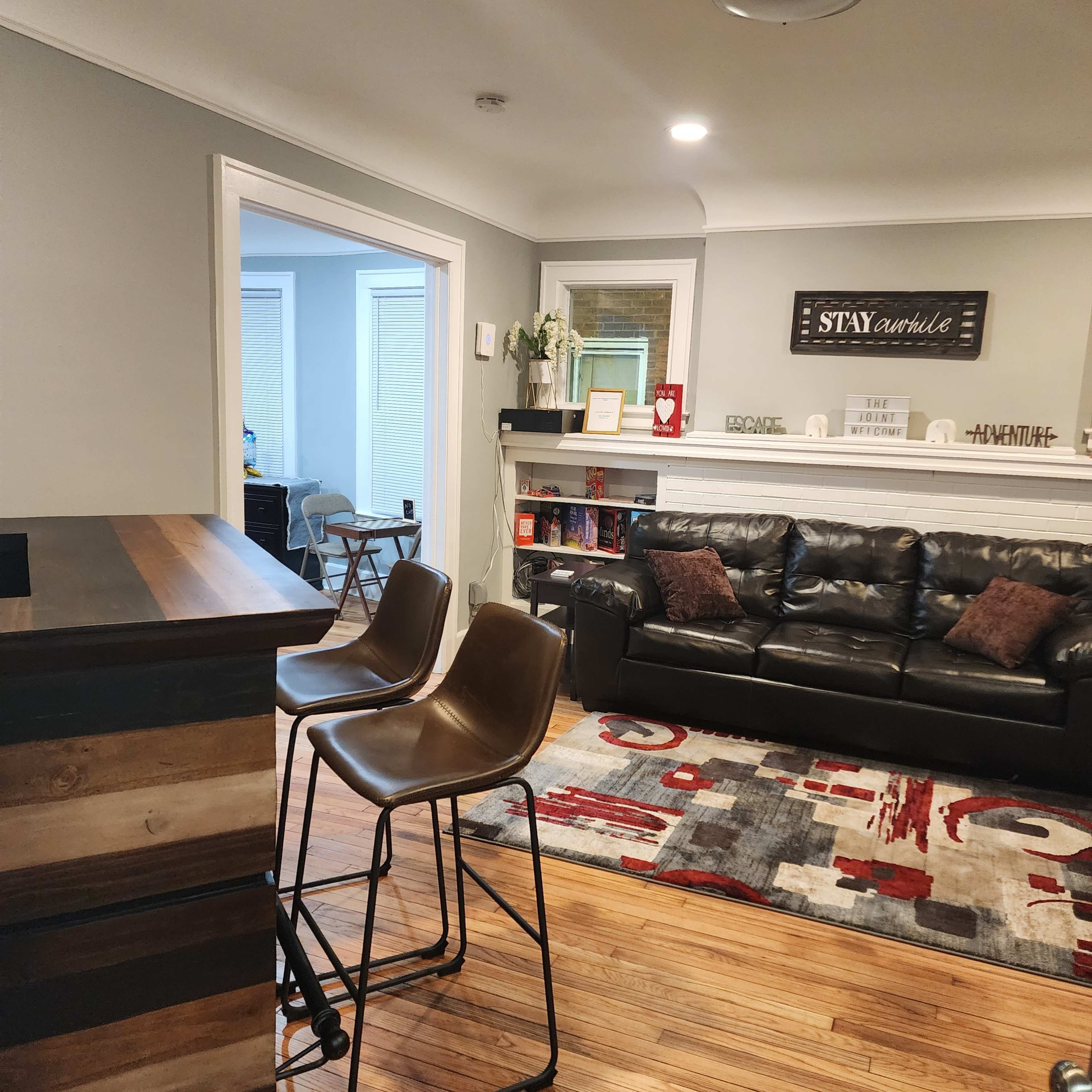 The image shows a living room with a black leather couch, a wooden bar counter with two stools, and a patterned area rug, featuring a bookshelf and a sign on the wall.