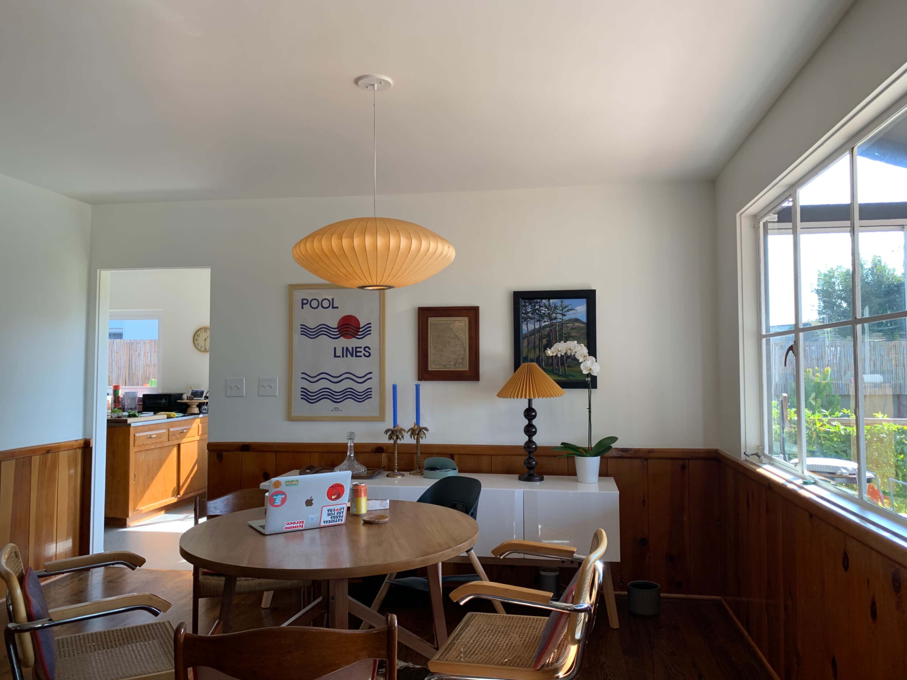 A dining area features a round wooden table surrounded by chairs, a pendant light overhead, and a large window with natural light.