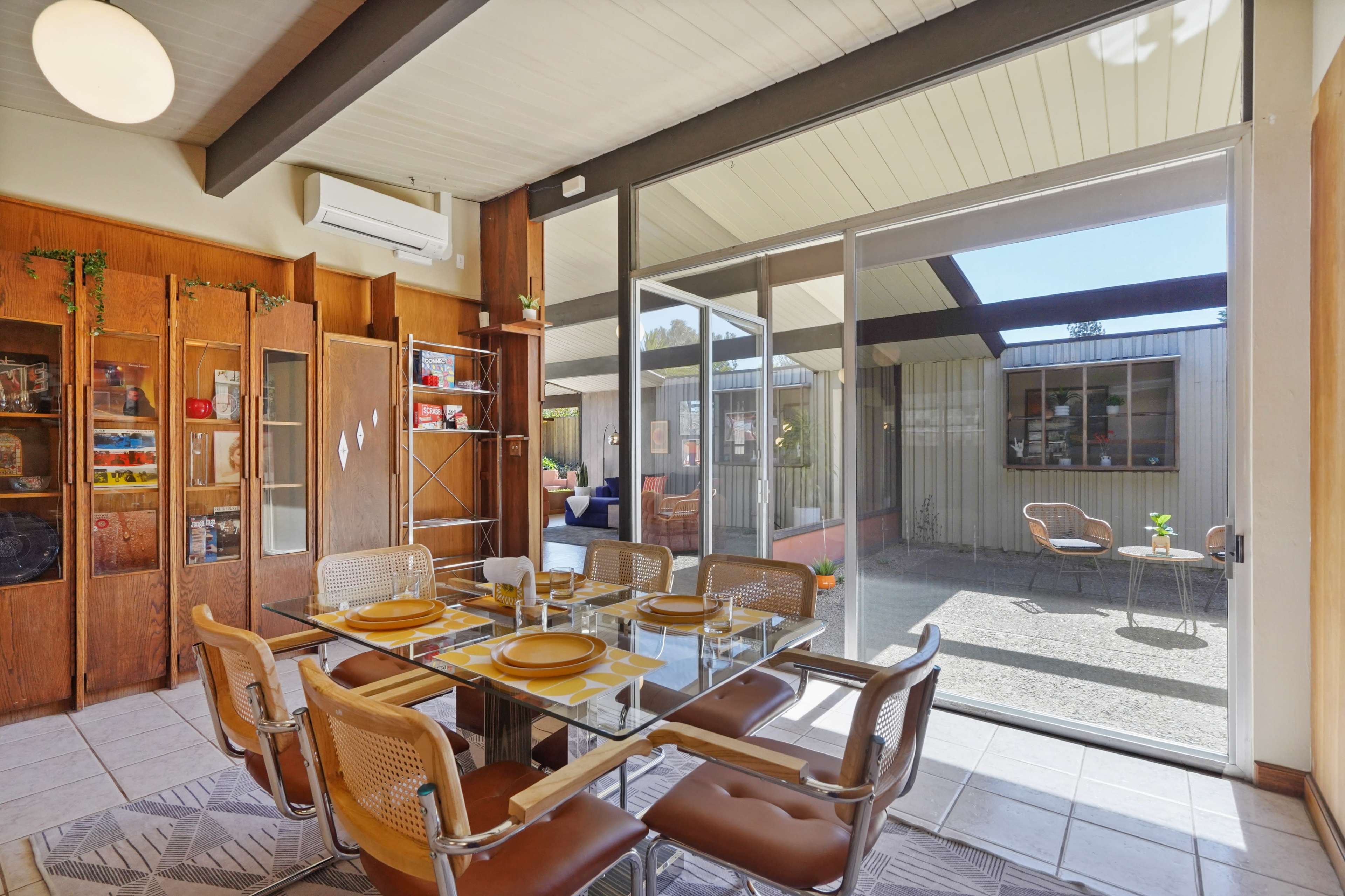 The image shows a dining area with a glass table and wooden cabinets, leading to an outdoor space visible through large sliding glass doors.