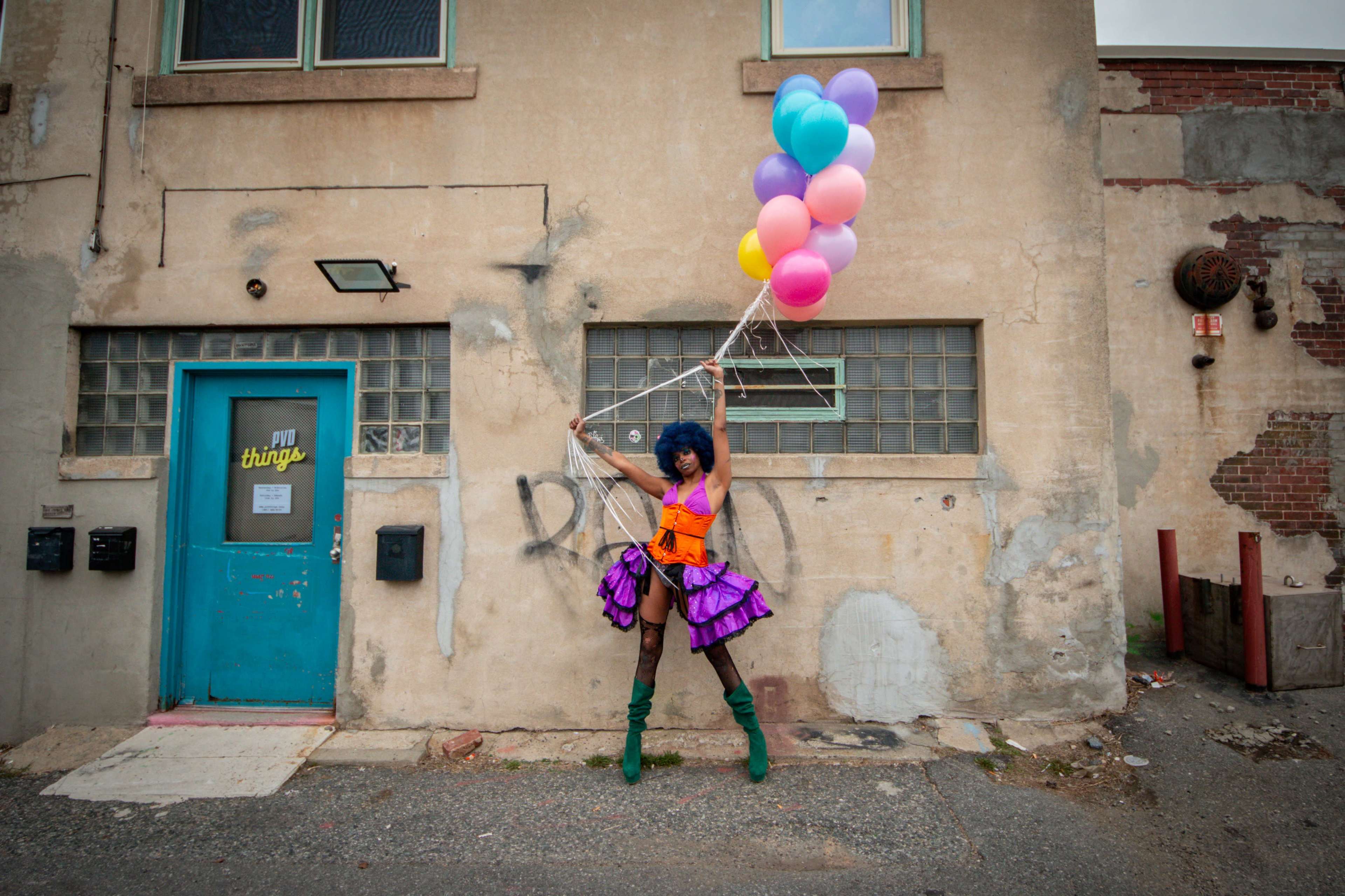 A person in a colorful outfit holds a cluster of balloons in front of an aged building with a blue door and graffiti.