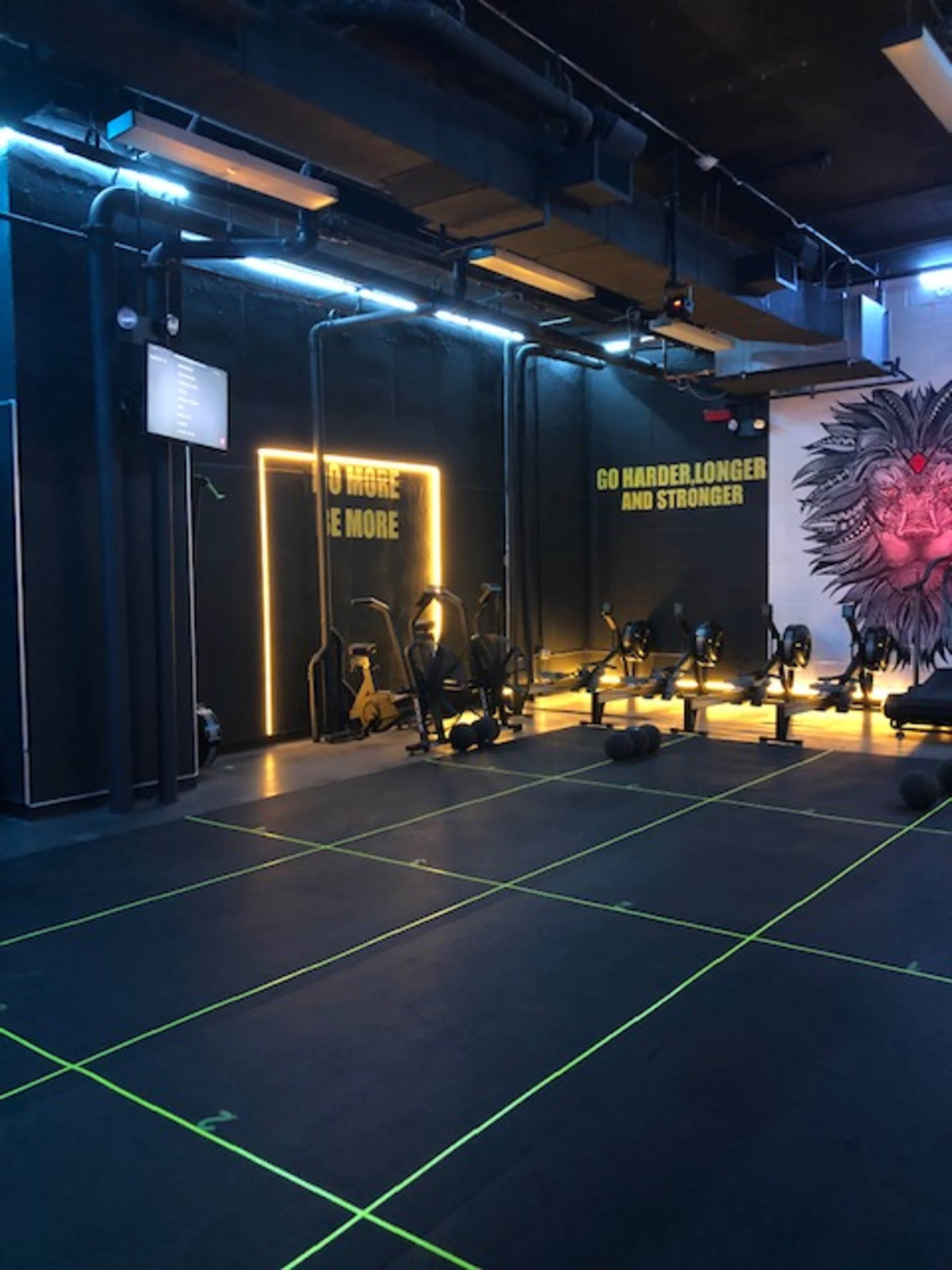 The image shows a modern gym interior with various exercise equipment arranged in a grid pattern, neon lighting, and motivational wall art.