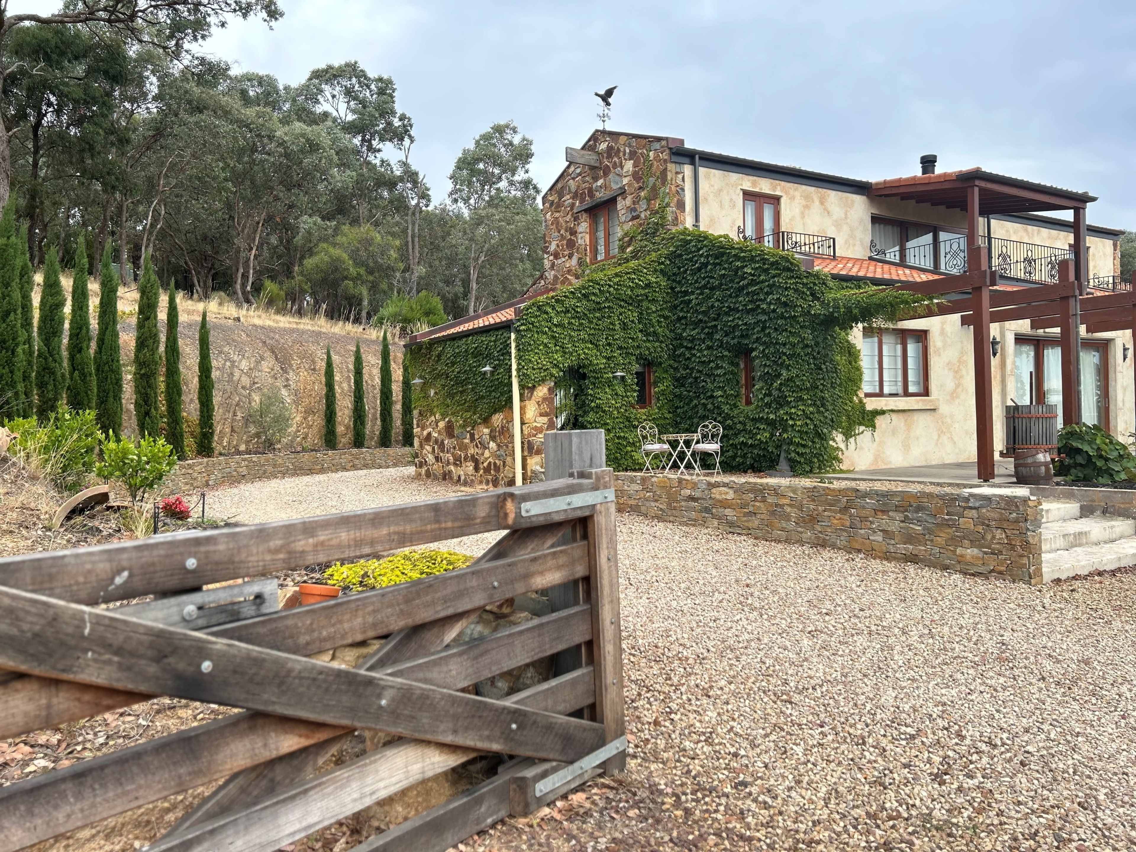 A stone and stucco house covered in greenery sits at the end of a gravel driveway, surrounded by tall cypress trees and rolling hills.