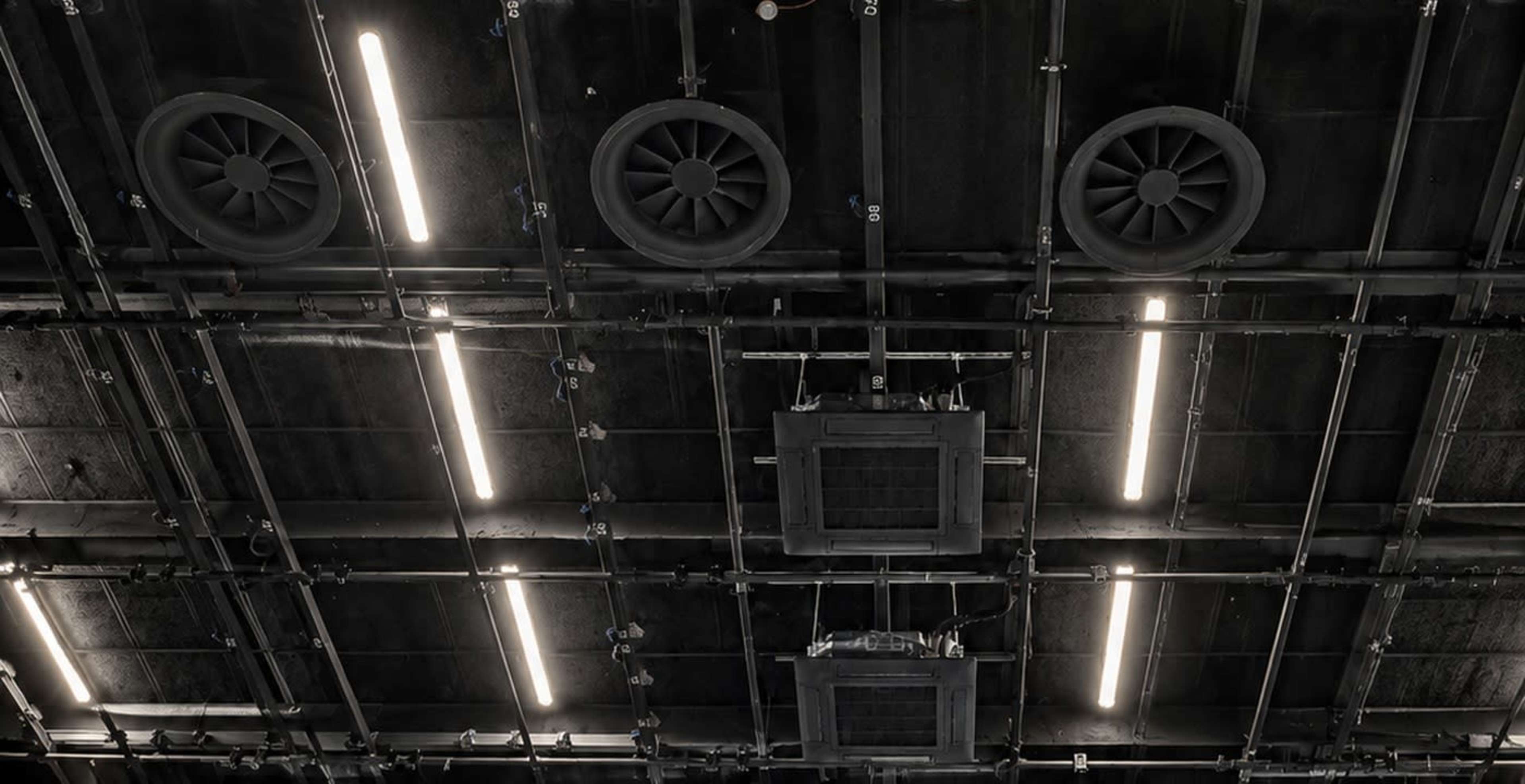 The image shows a black ceiling with industrial fans and elongated light fixtures.