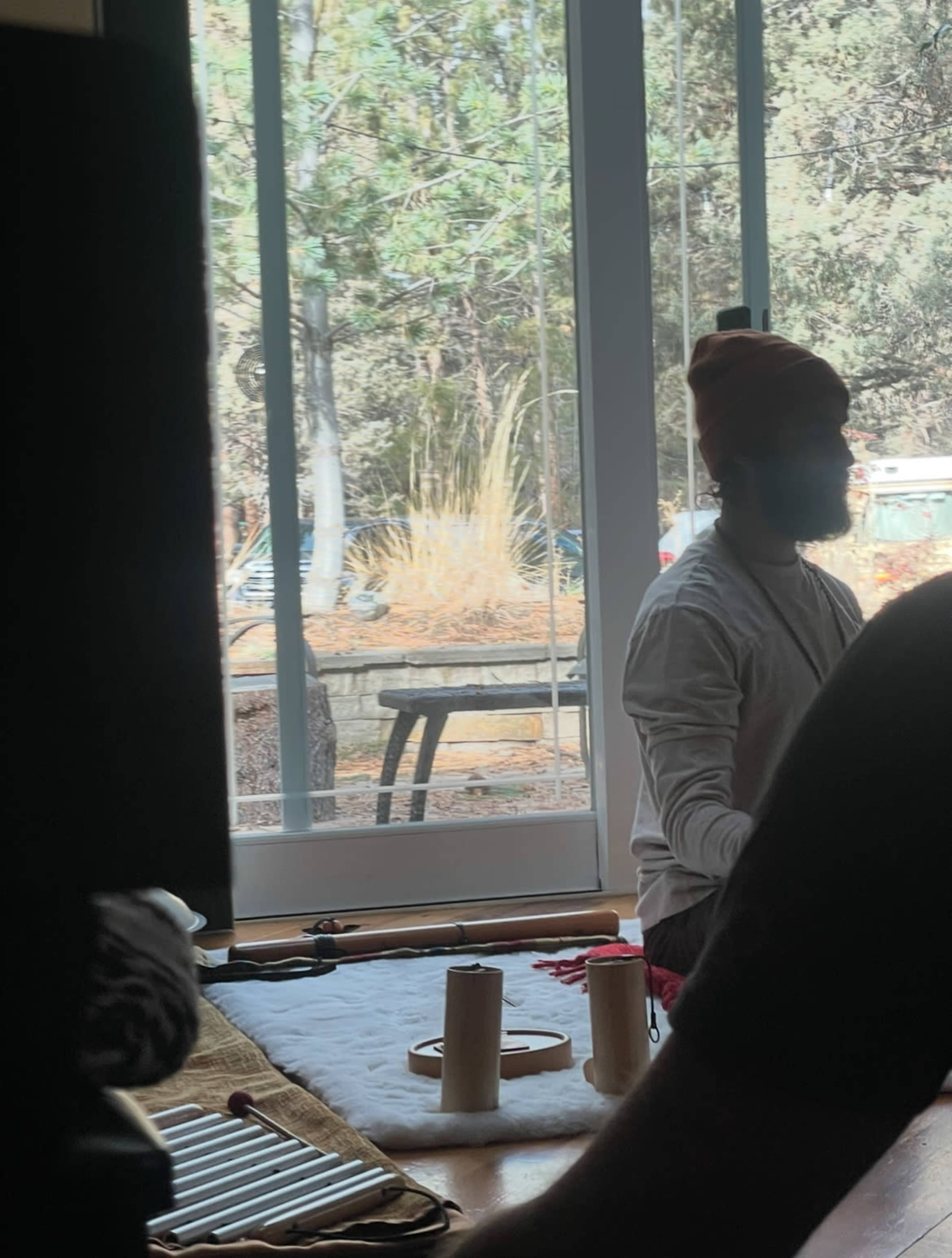 A person wearing a beanie sits beside a wooden table with a view of trees outside through a large window.