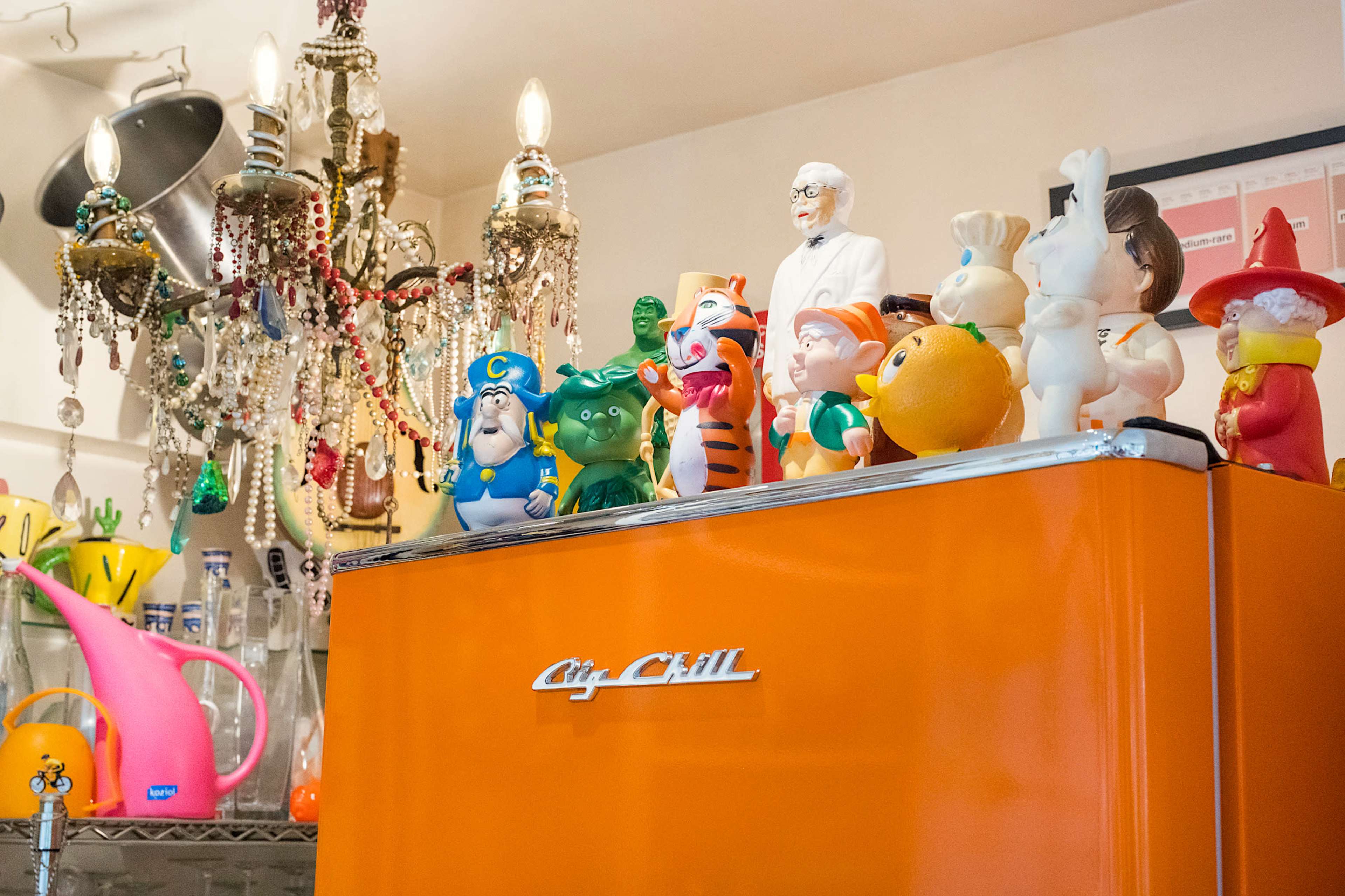 An orange refrigerator is adorned with a collection of colorful plastic figurines, while vintage chandeliers and decorative items hang above.
