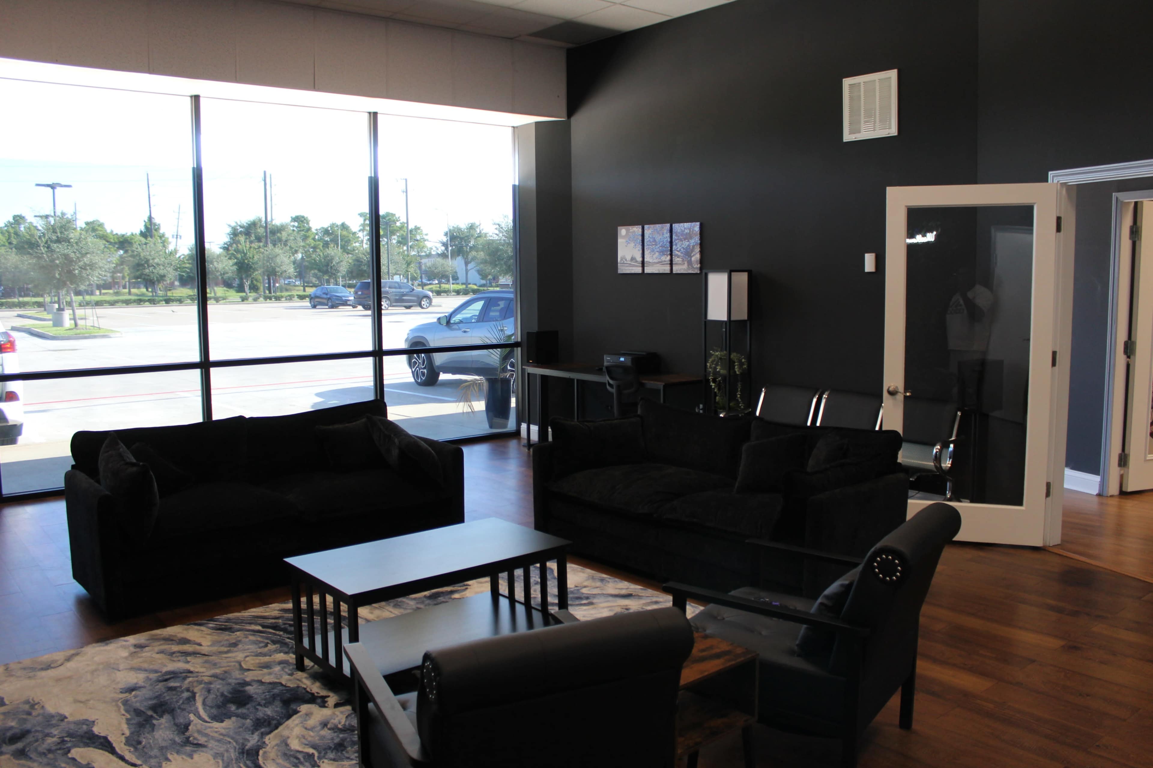 The image shows a spacious indoor seating area with black sofas, a coffee table, and large windows allowing natural light to enter.