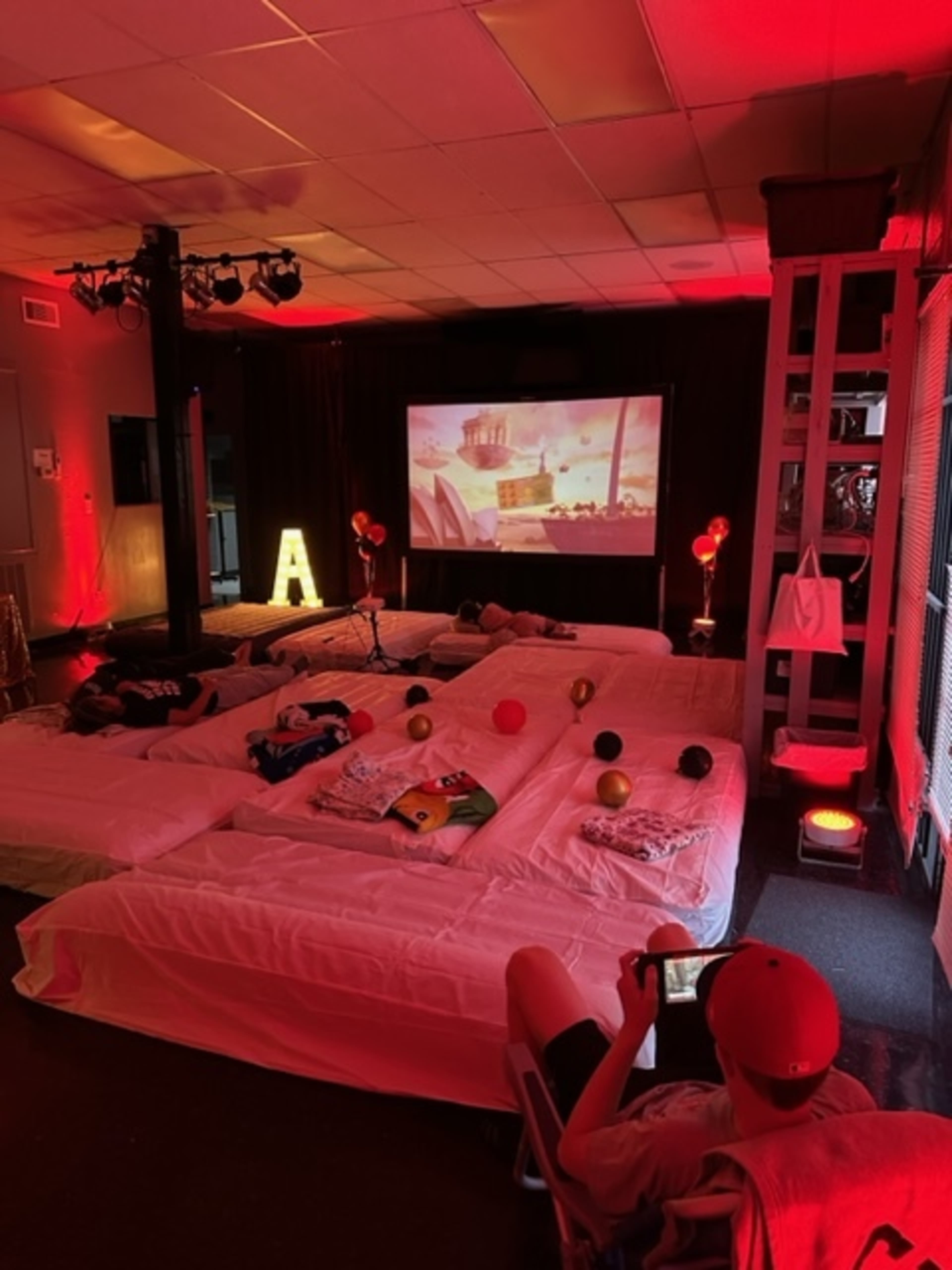 The scene shows a cozy movie setup with white mattresses arranged on the floor, a projector displaying a film on a large screen, and balloons decorating the space.