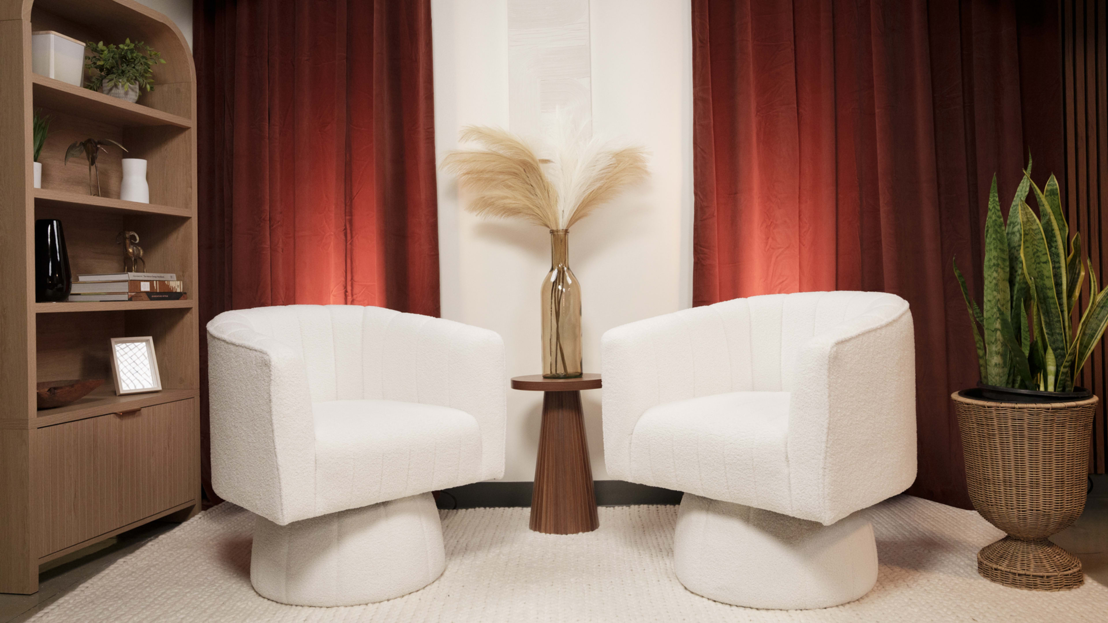 The image shows a cozy interior setting with two white upholstered chairs facing each other, a small wooden side table between them, and a decorative vase with tall dried grass in the center, all framed by warm red curtains.