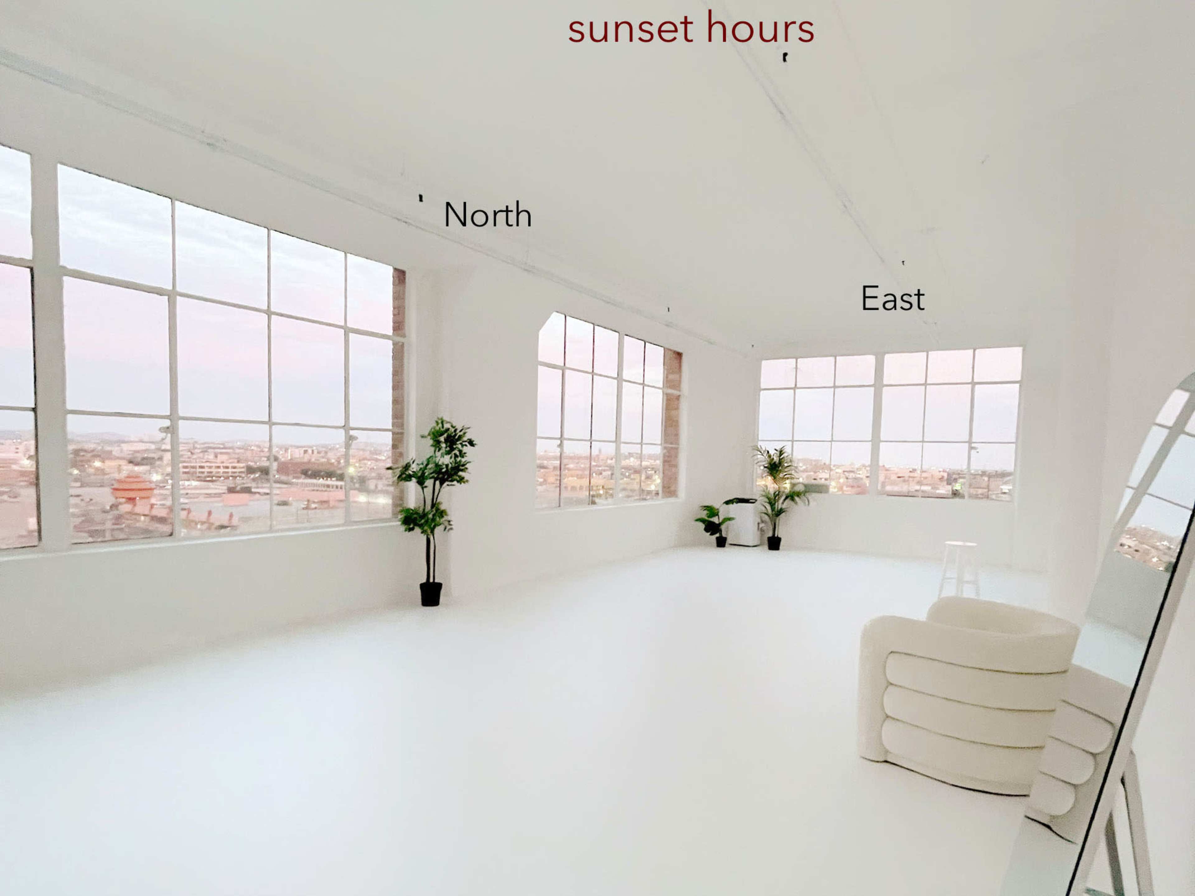 A bright, minimalist studio features large windows with views of a cityscape, labeled with cardinal directions.