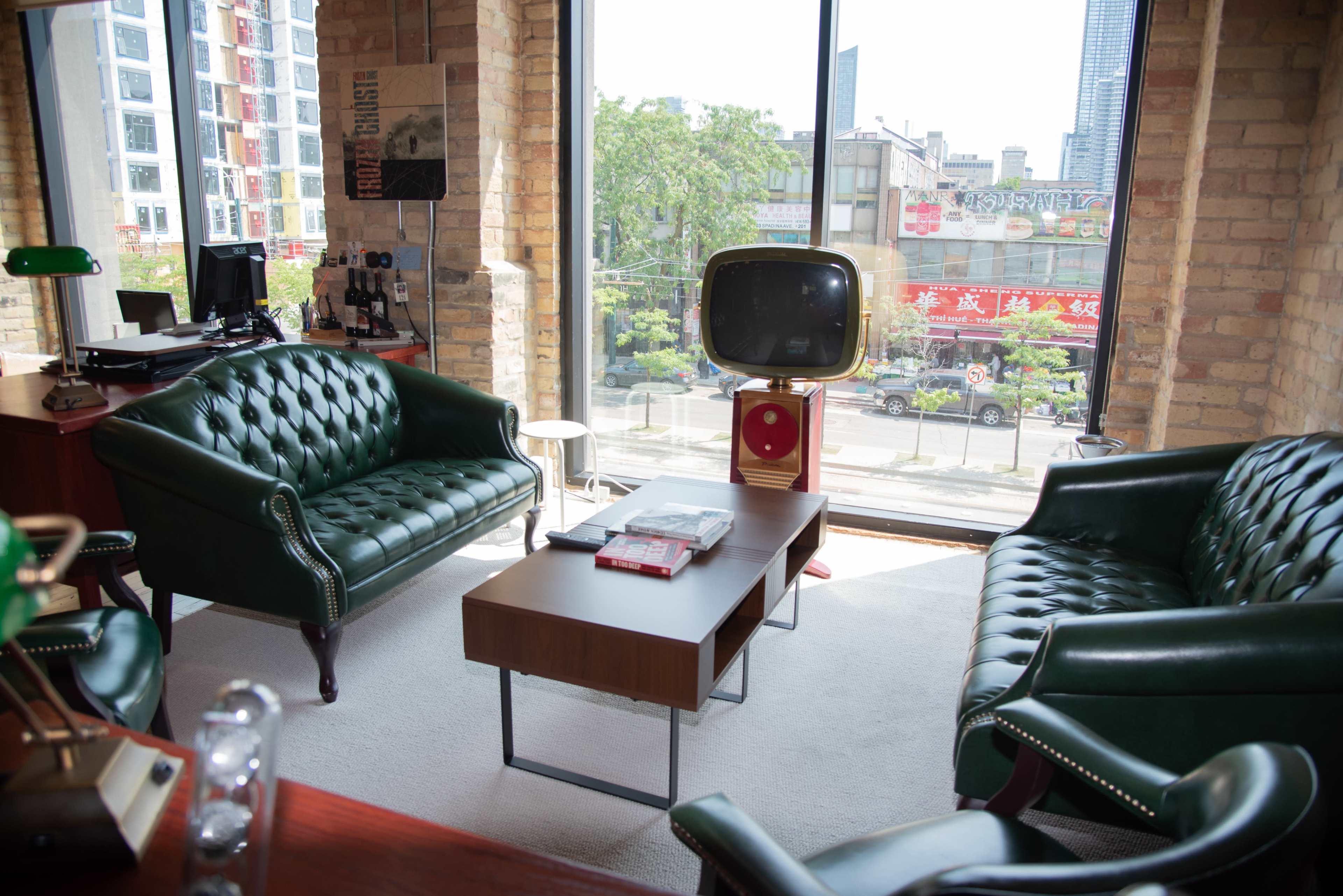 The image shows a modern living room with two green leather sofas, a wooden coffee table, and a vintage television set positioned near a large window that offers a view of a city street.