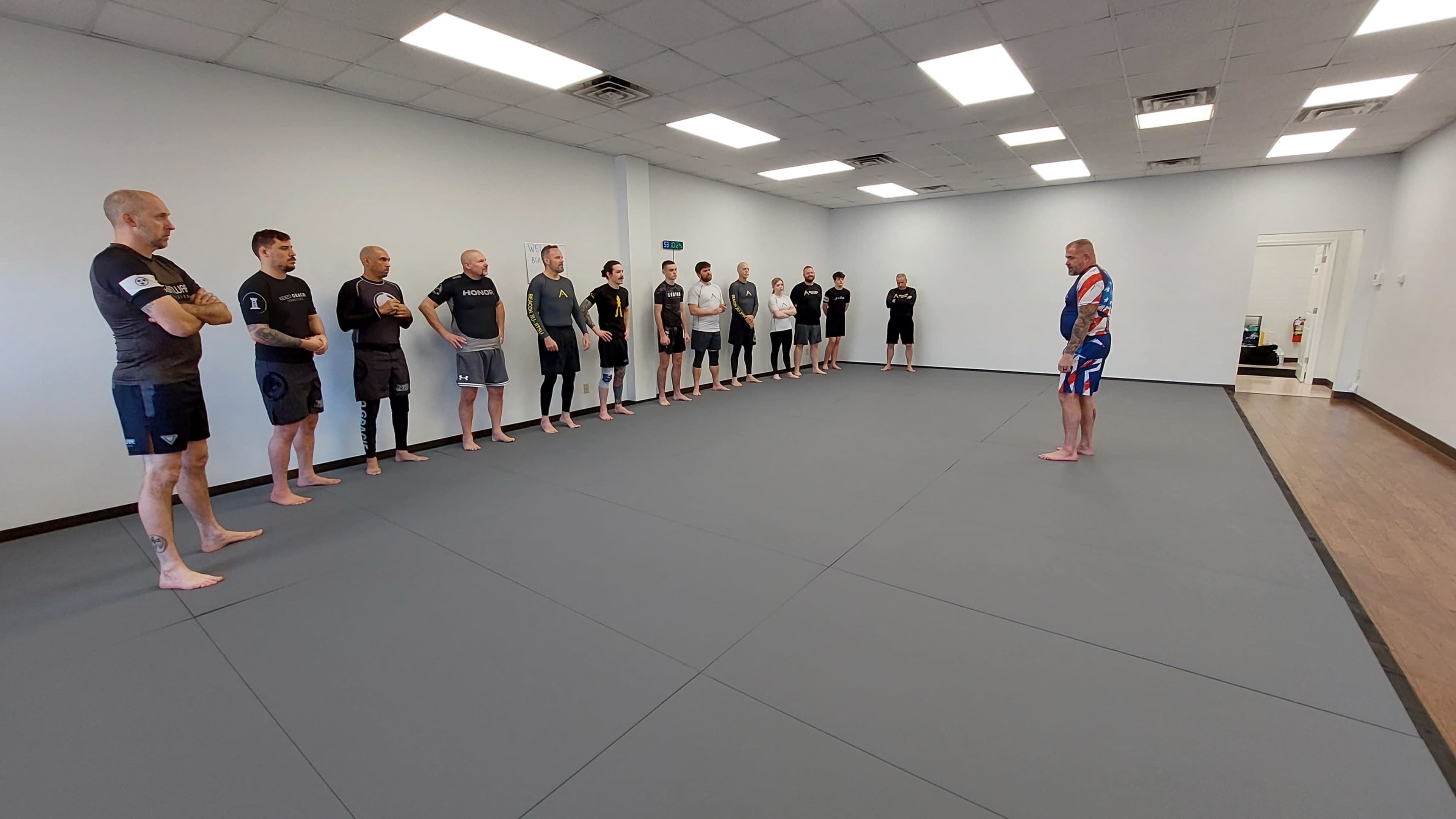 A group of men stands in two lines on a gray mat in a spacious training room, facing a coach who is addressing them.