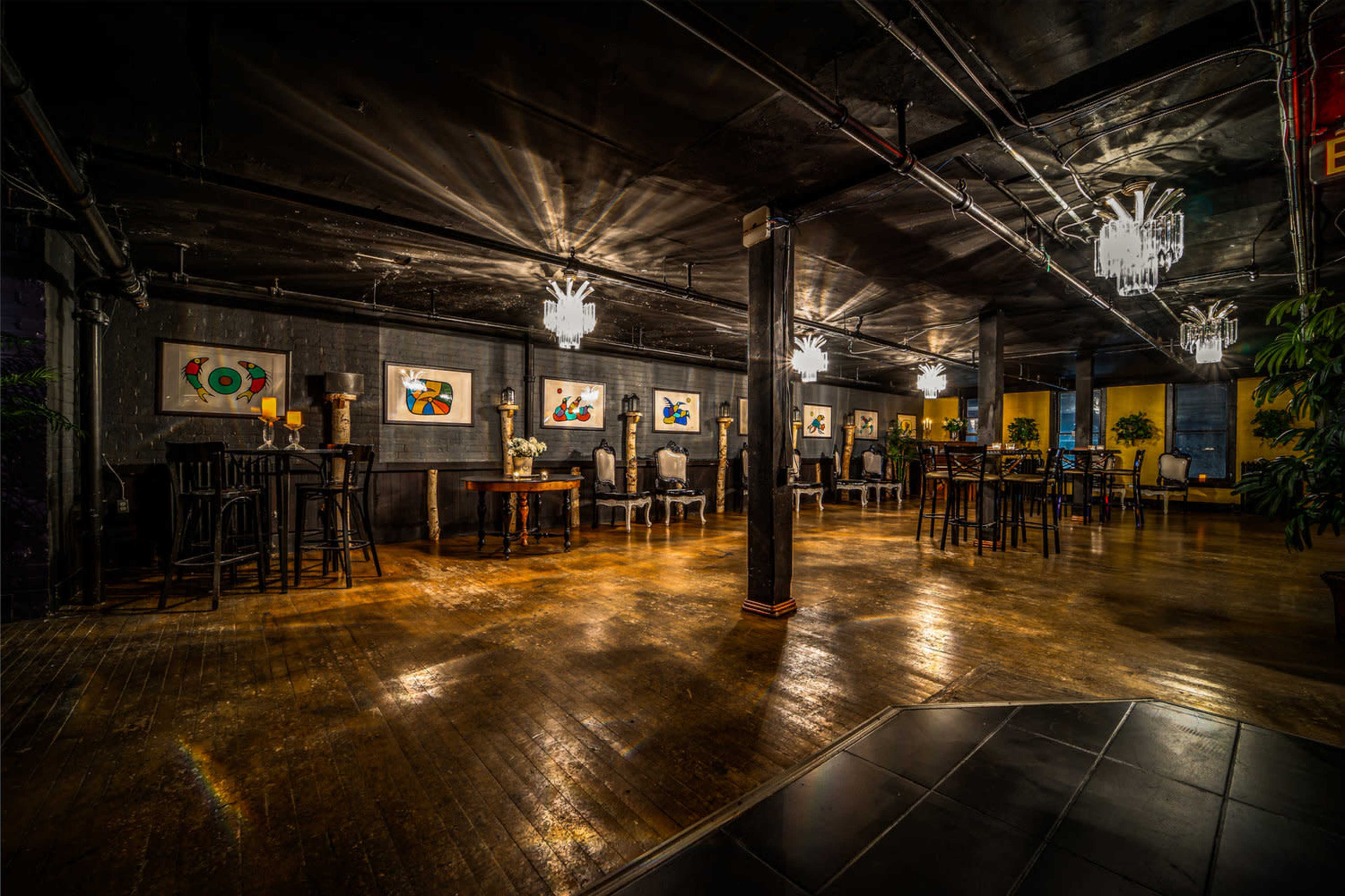 A dimly lit event space with wooden flooring, adorned with framed artwork on the walls and various seating arrangements.