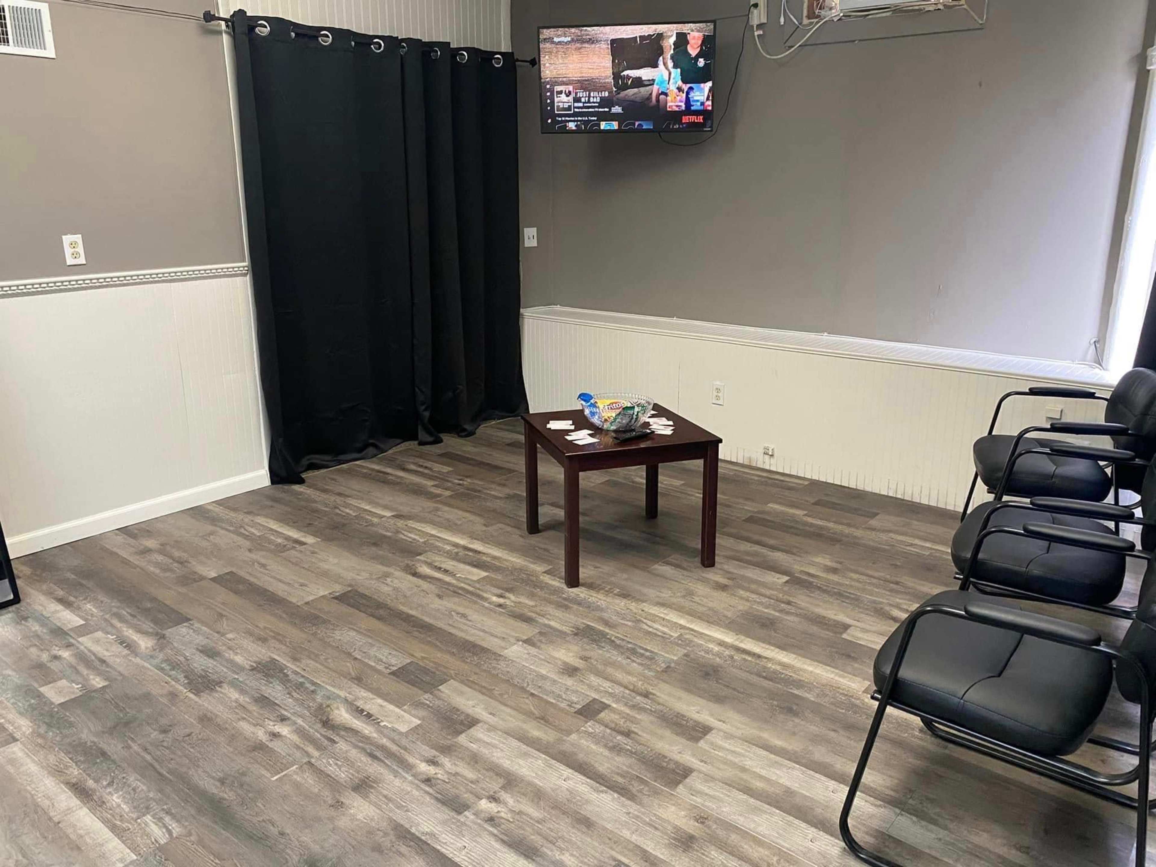 A waiting room features a small table in the center, black chairs along one side, and a television mounted on the wall.