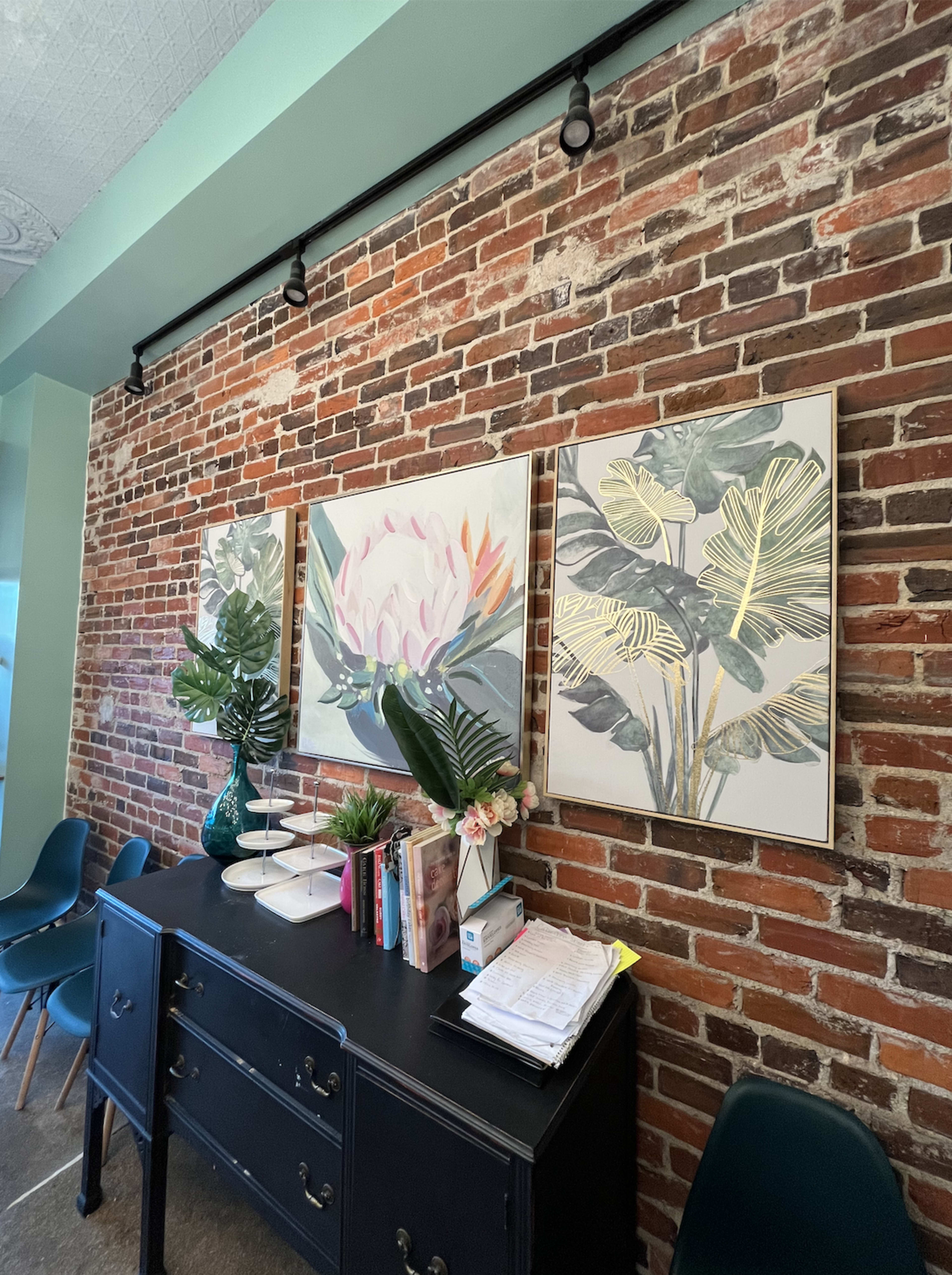 A black dresser against a brick wall adorned with three framed botanical prints and decorative plants.
