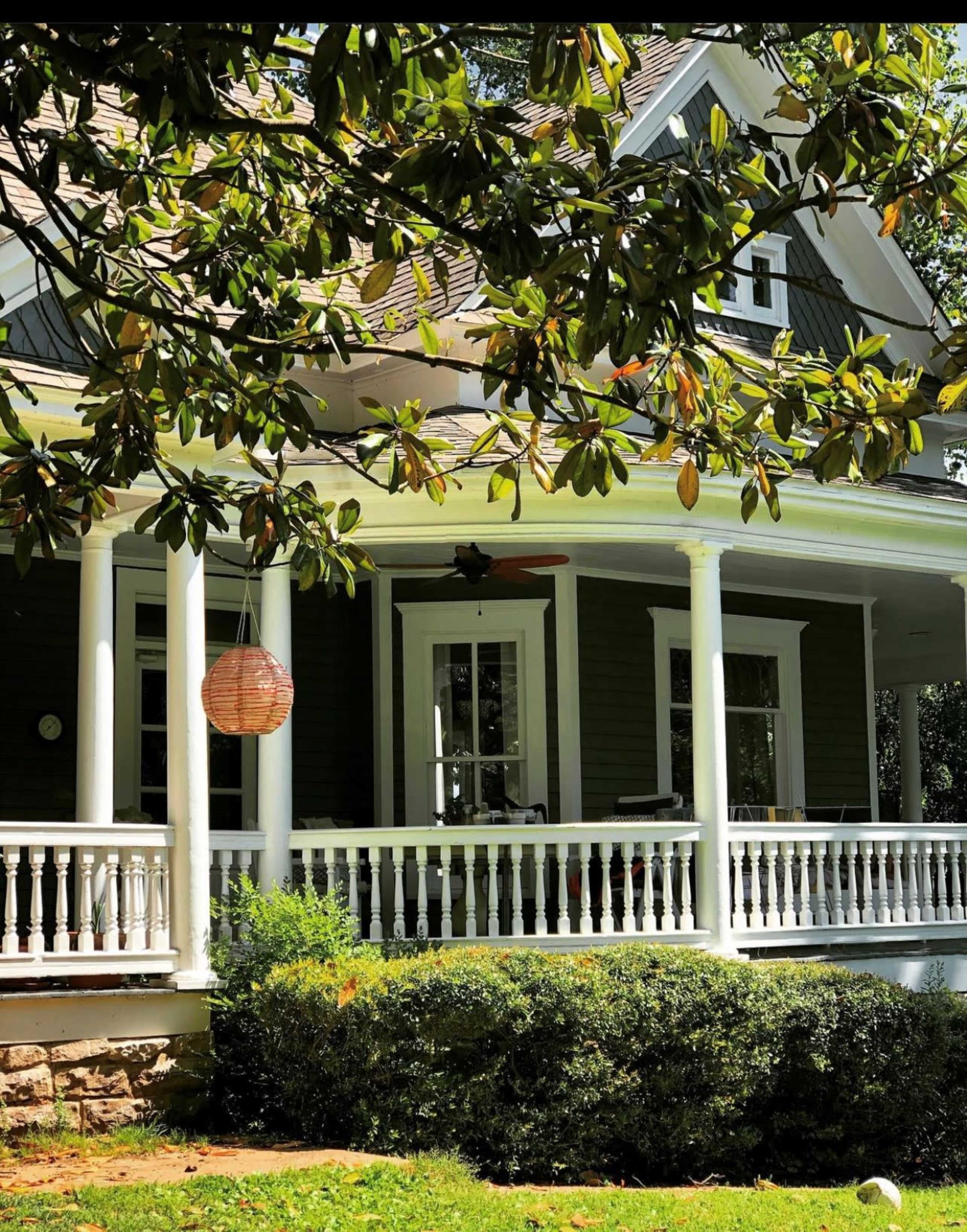 A large, two-story house with a wraparound porch is partially shaded by a magnolia tree.