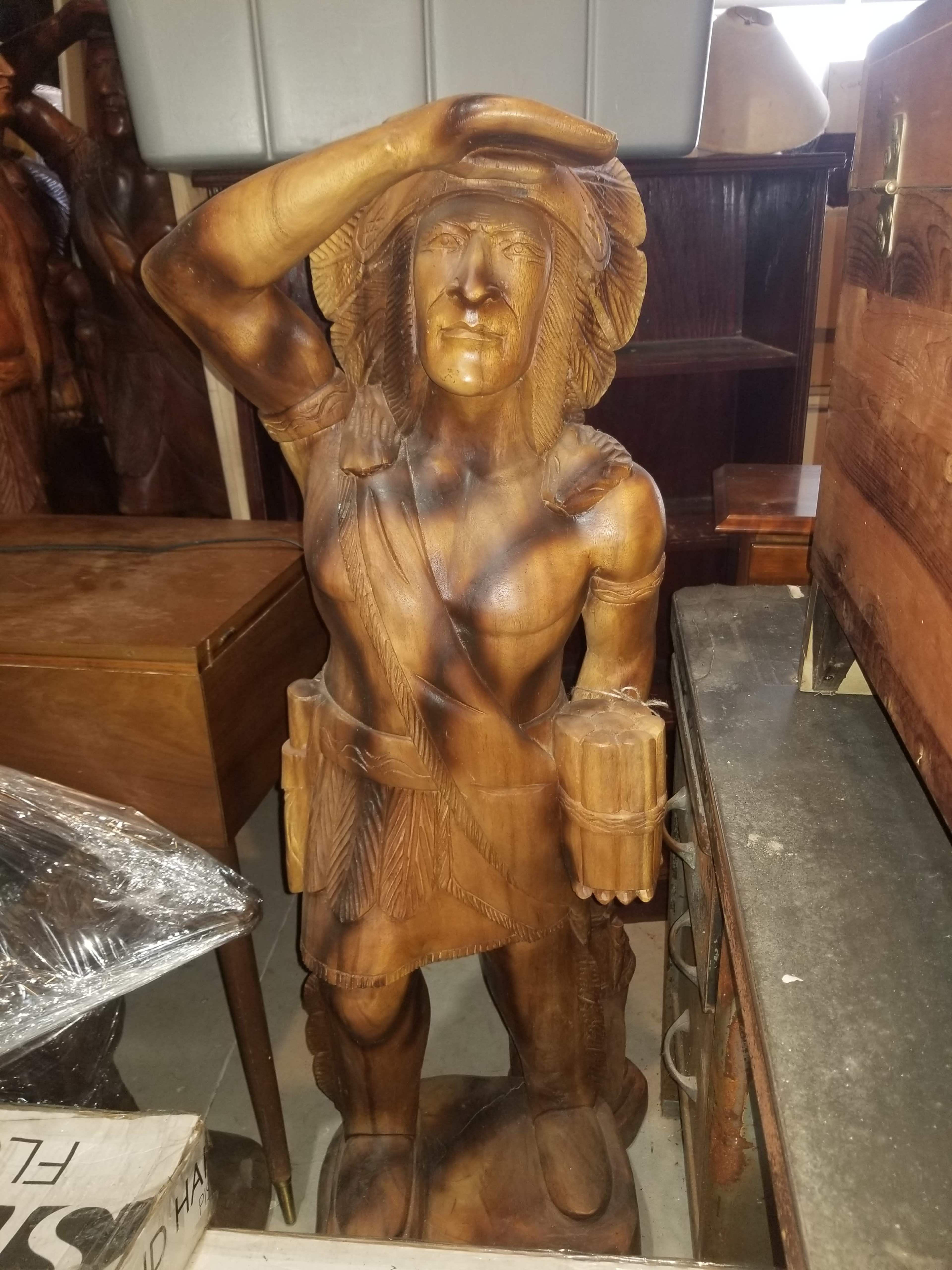 A wooden statue of a figure with one hand raised to its forehead and holding a cylinder in the other hand, surrounded by various items in a cluttered space.
