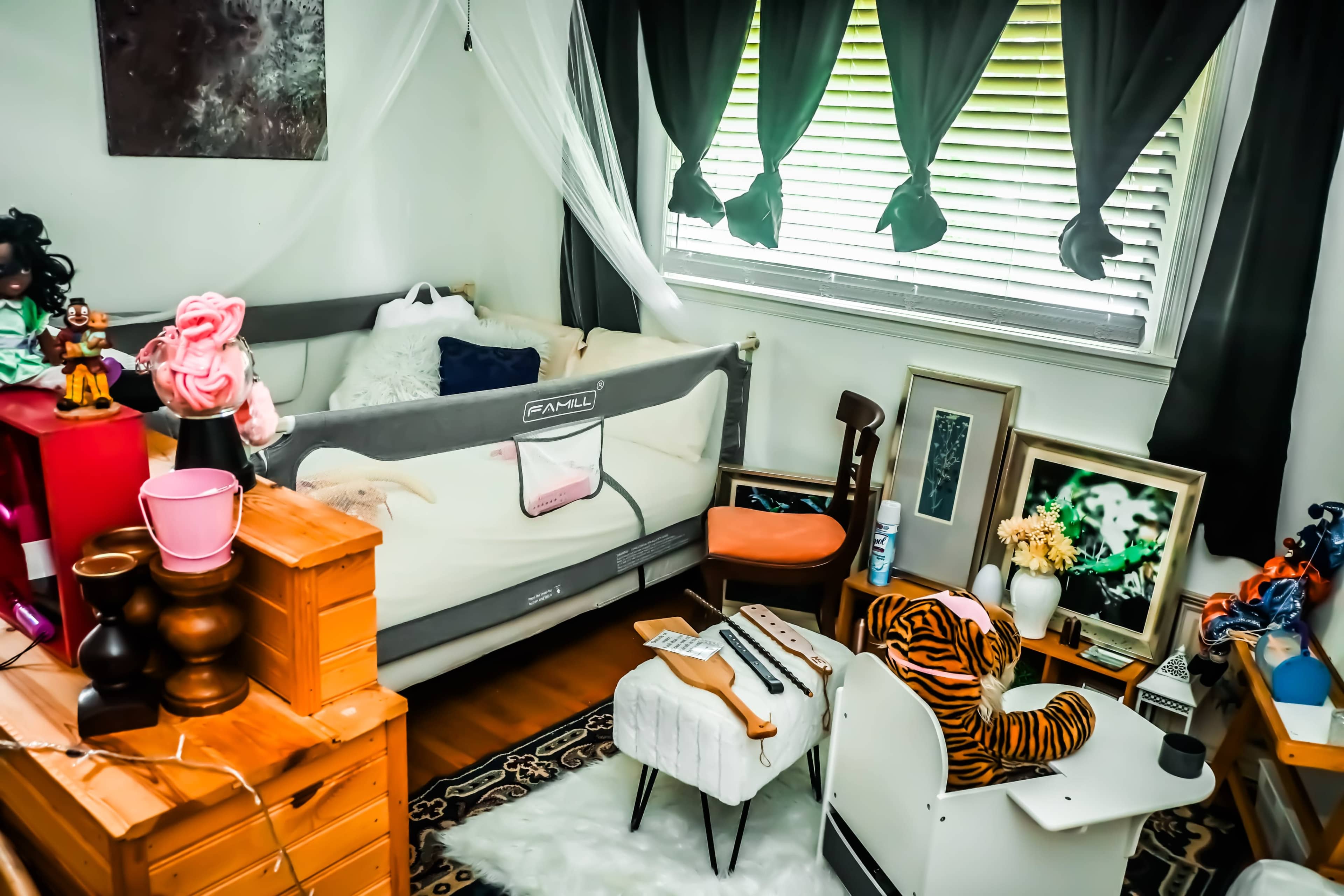 The image shows a small bedroom with a gray bed, wooden furniture, decorative items, and a window draped with black curtains.