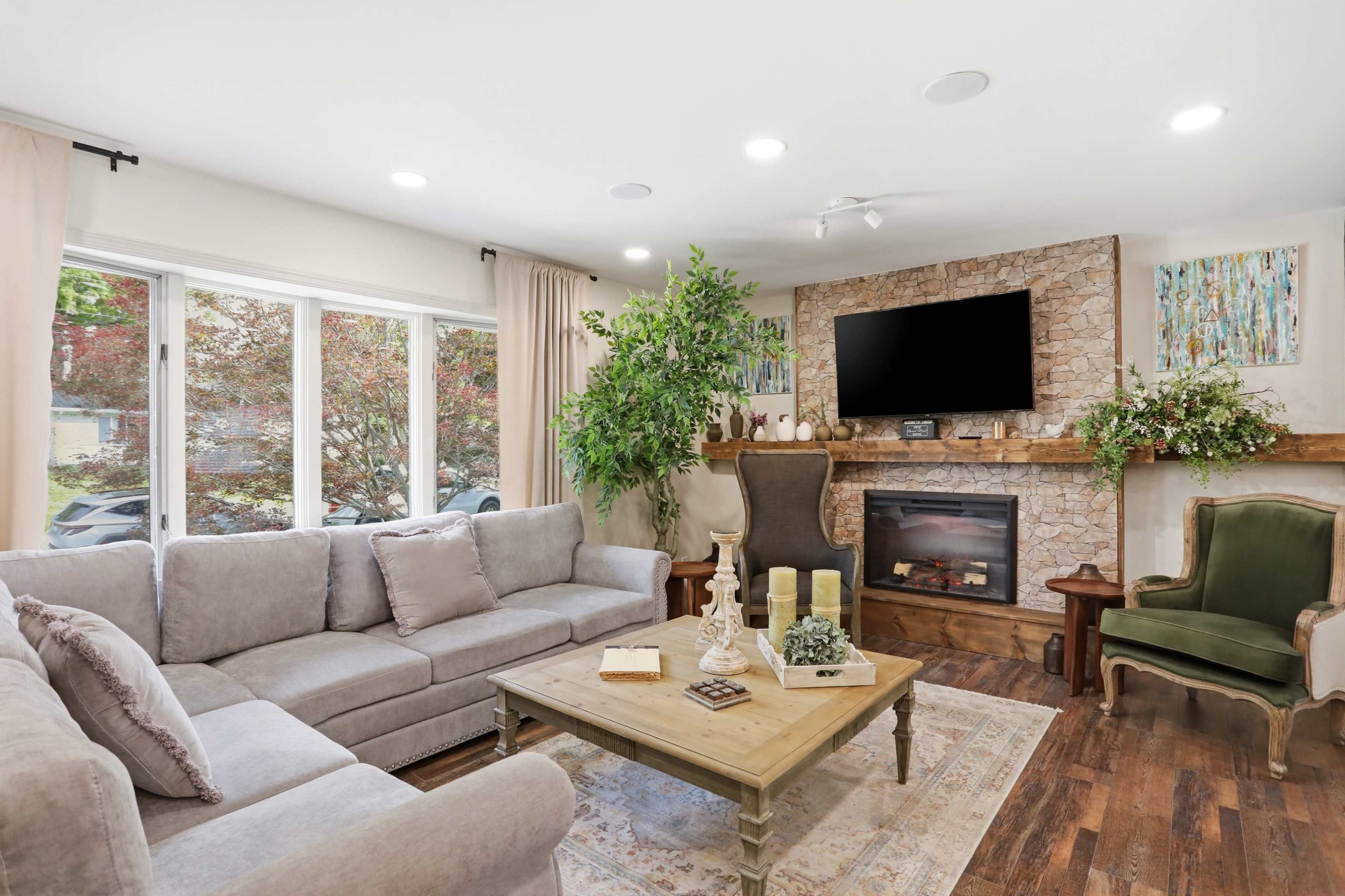 The living room features a gray sectional sofa, a wooden coffee table, an electric fireplace, and large windows that allow natural light to flow in.