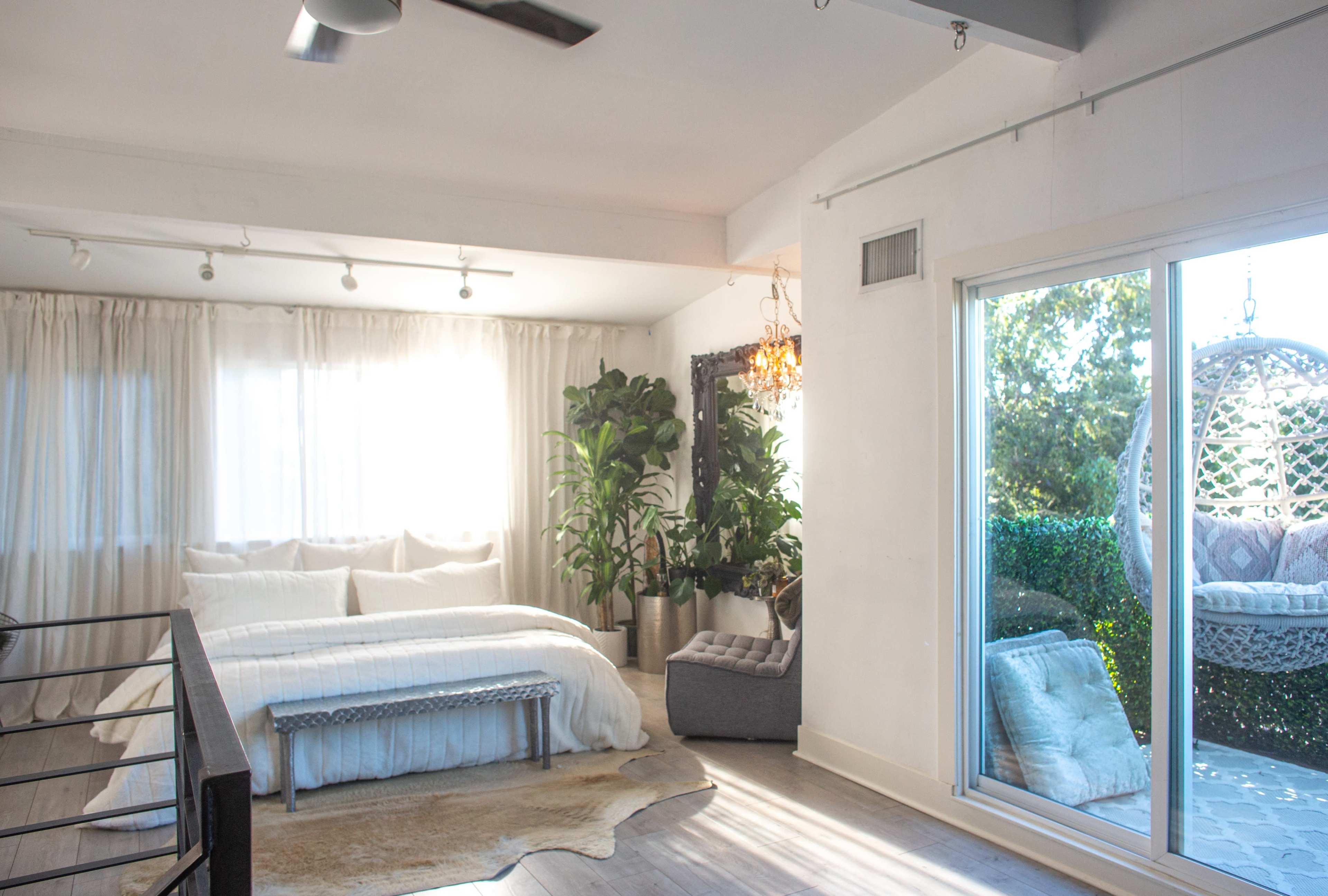 A bright bedroom with a large bed, a decorative bench, indoor plants, and a sliding glass door leading to an outdoor space with a hanging chair.