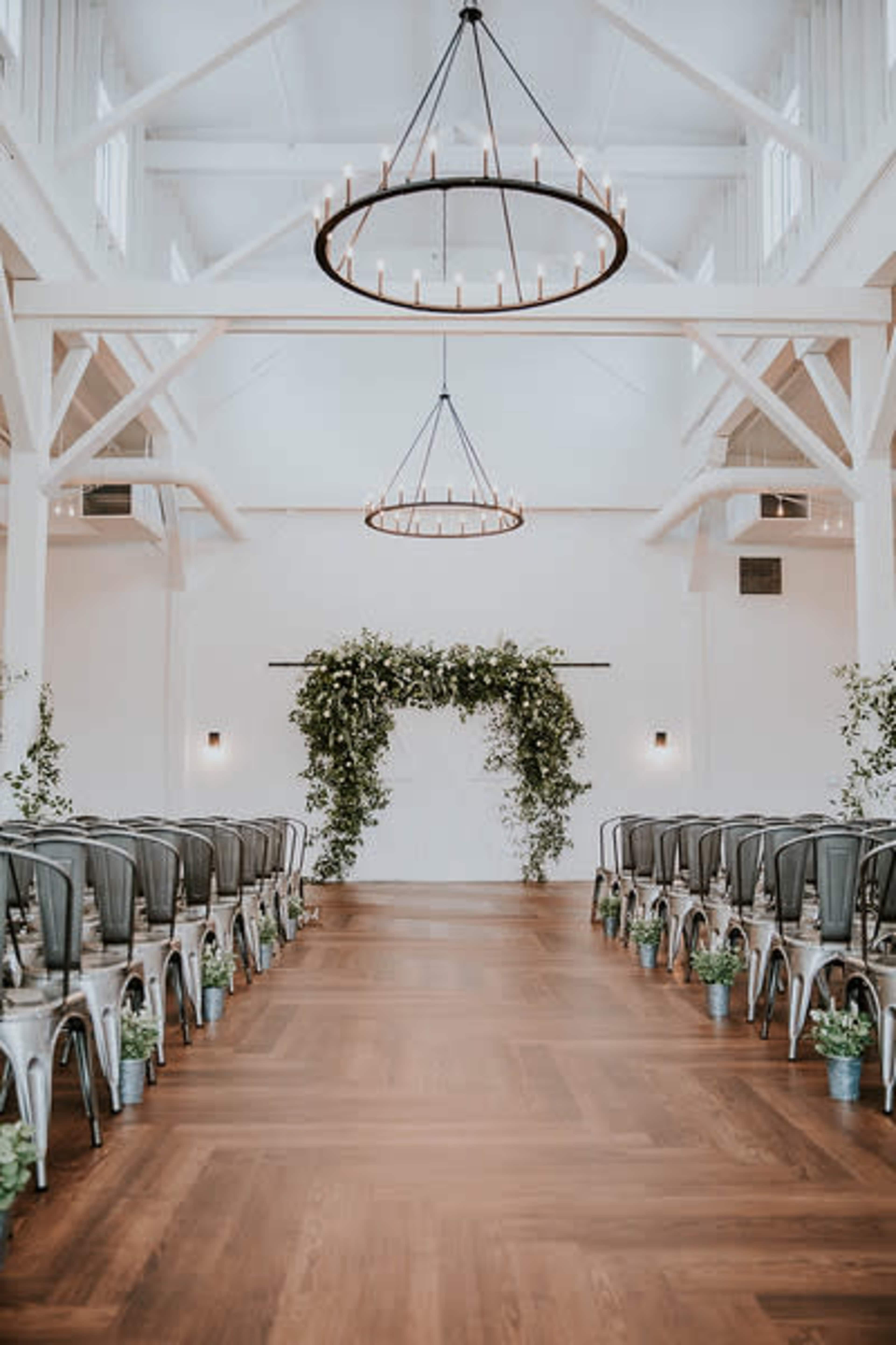 A spacious indoor venue is set up for a wedding ceremony, featuring rows of metal chairs facing a floral arch under two large, circular light fixtures.