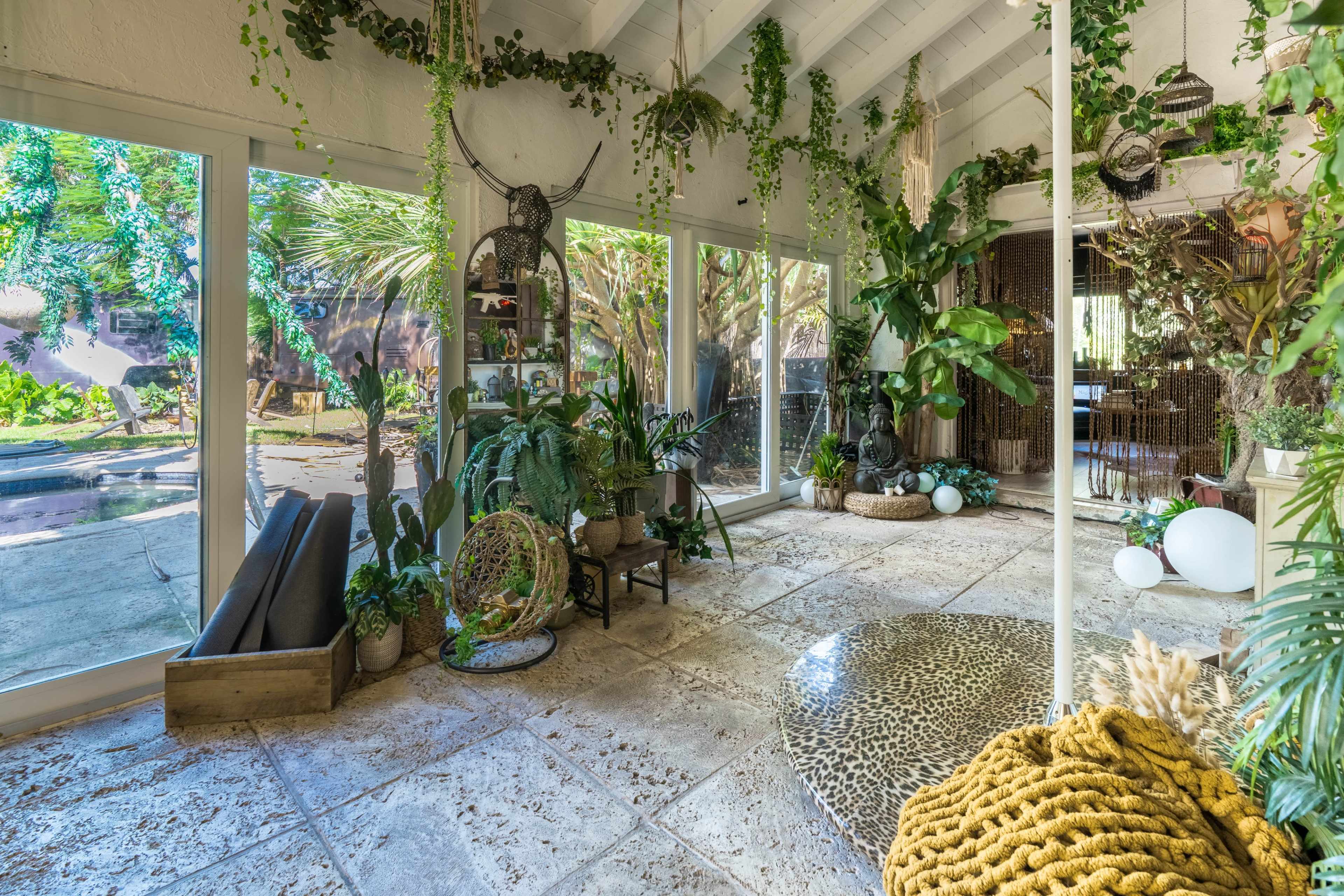 The image shows a spacious, well-lit interior filled with various plants and decorative elements, featuring large glass doors that open to a landscaped outdoor area.