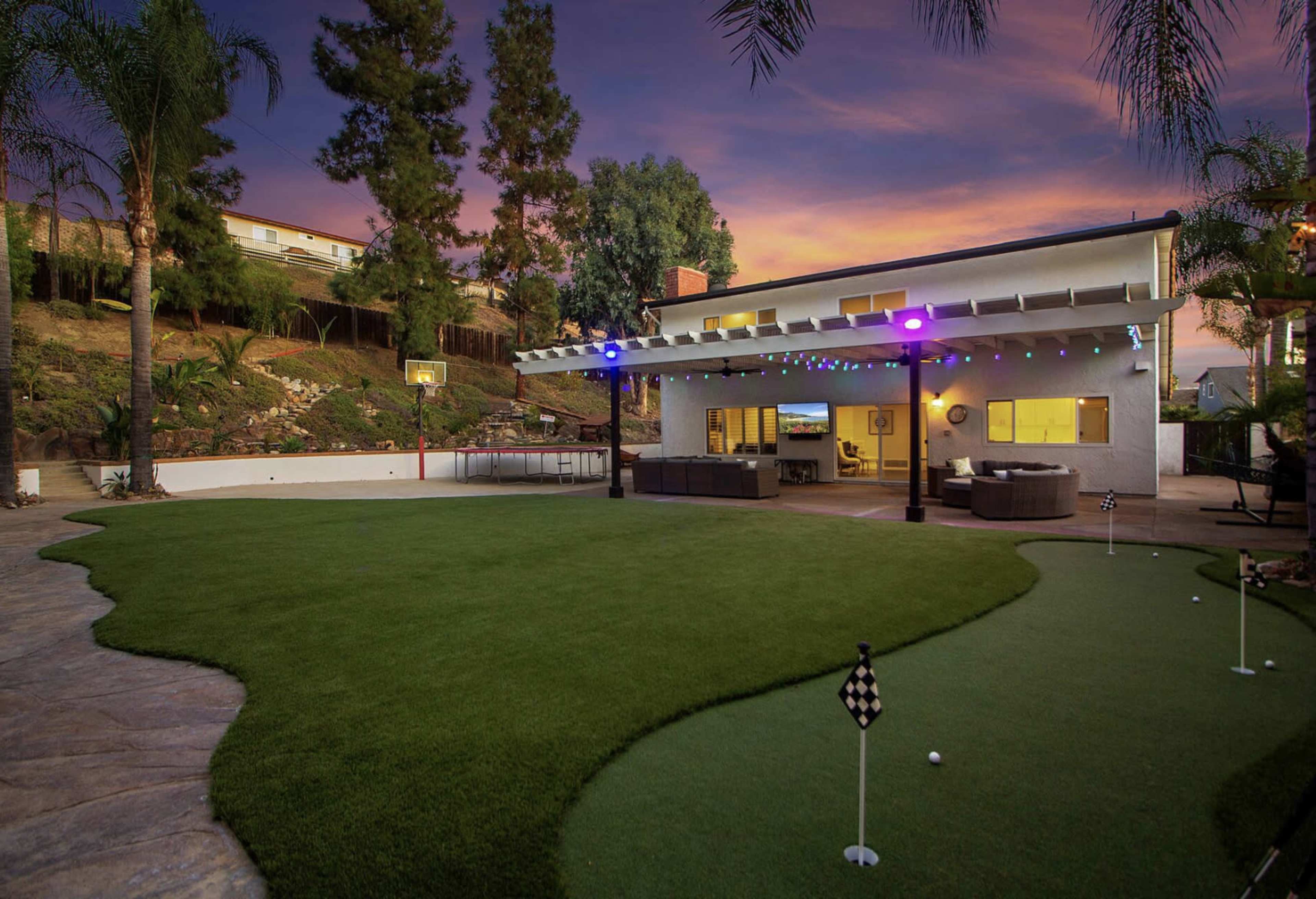 A backyard with a golf putting green, modern patio furniture, and a house in the background, all set against a colorful sunset.