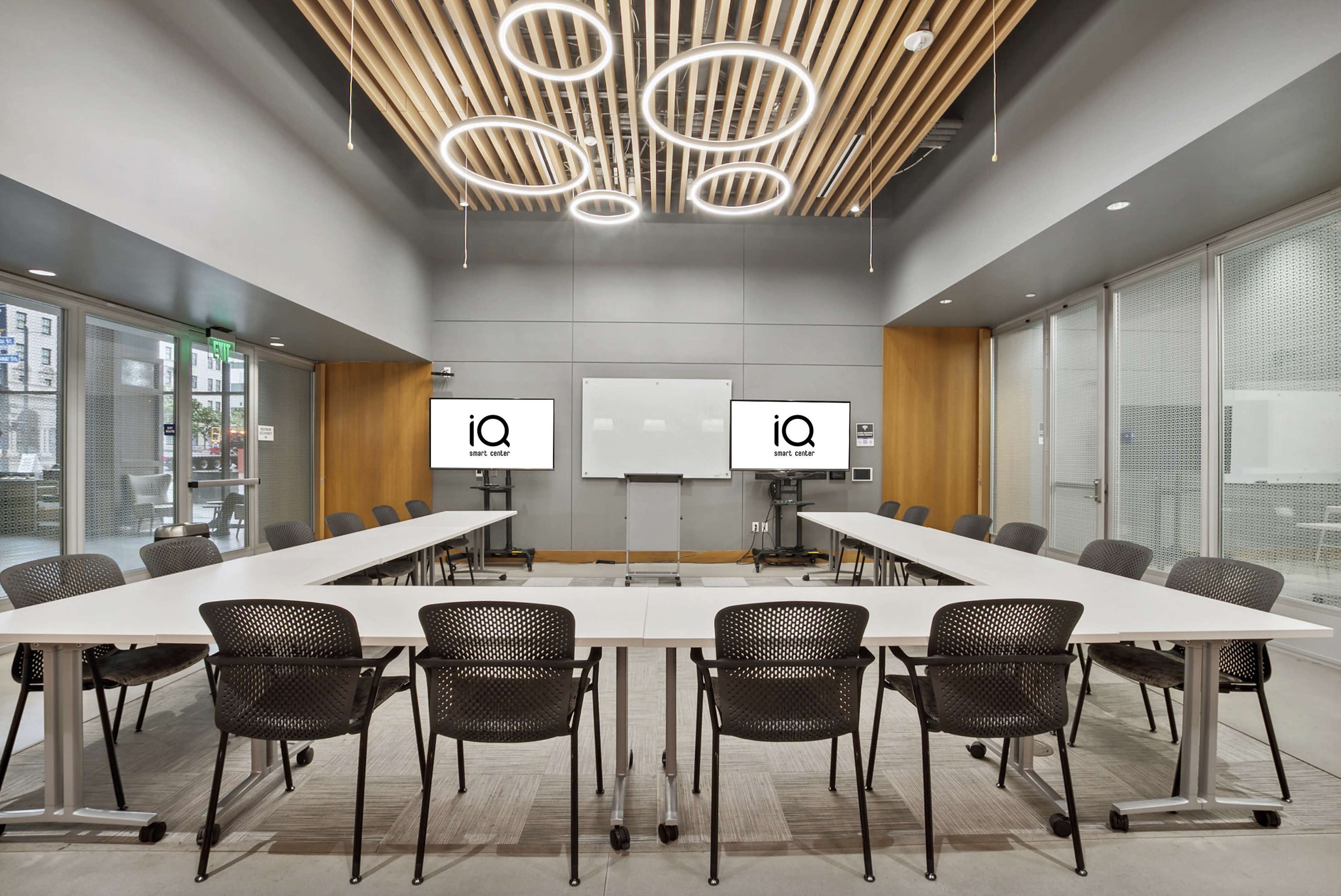 A modern conference room features a large oval table surrounded by black chairs, with two screens displaying the logo "iQ" and a whiteboard on the wall.