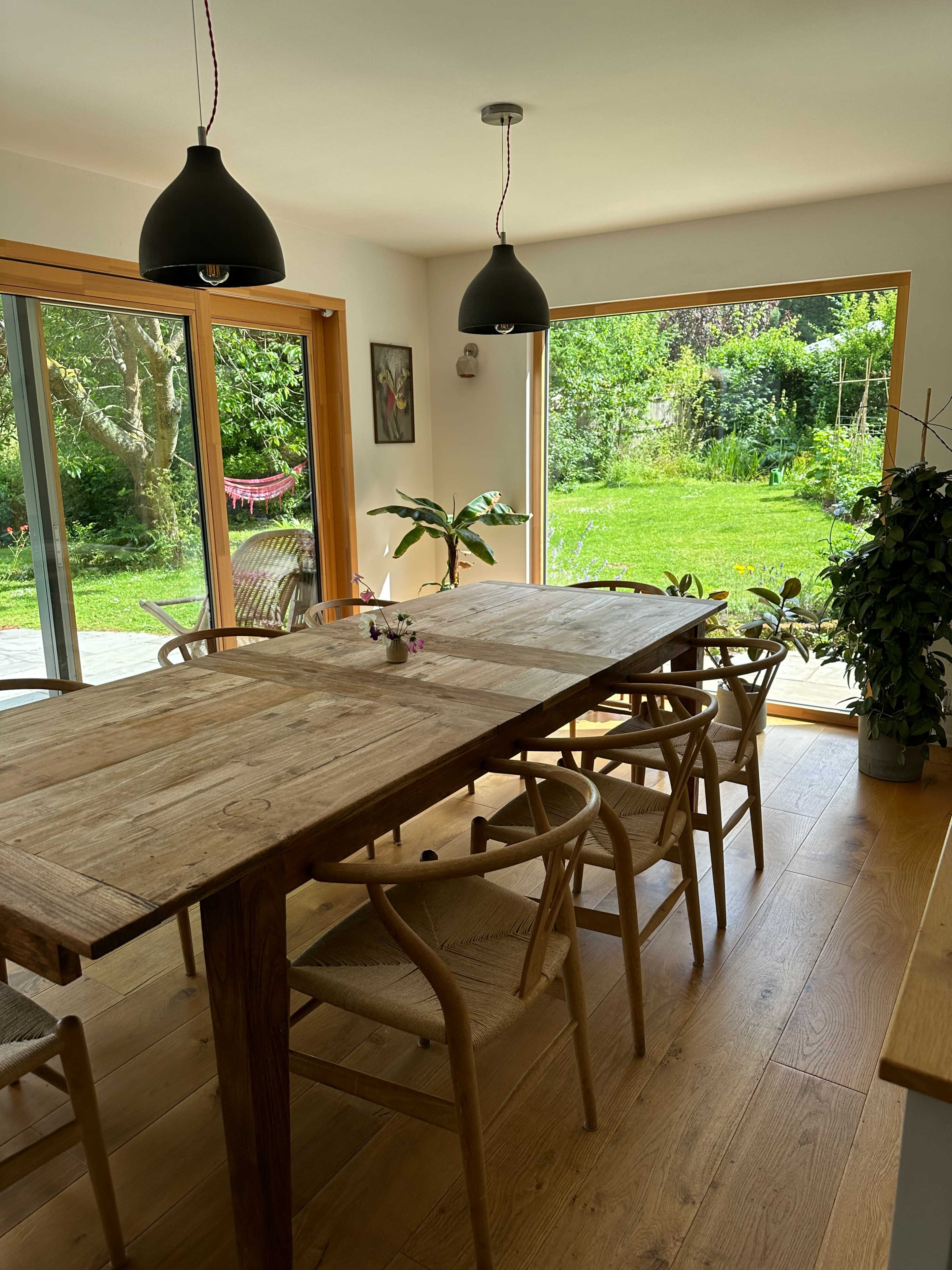 A spacious dining area featuring a long wooden table, surrounded by chairs, with large glass doors opening to a green garden.