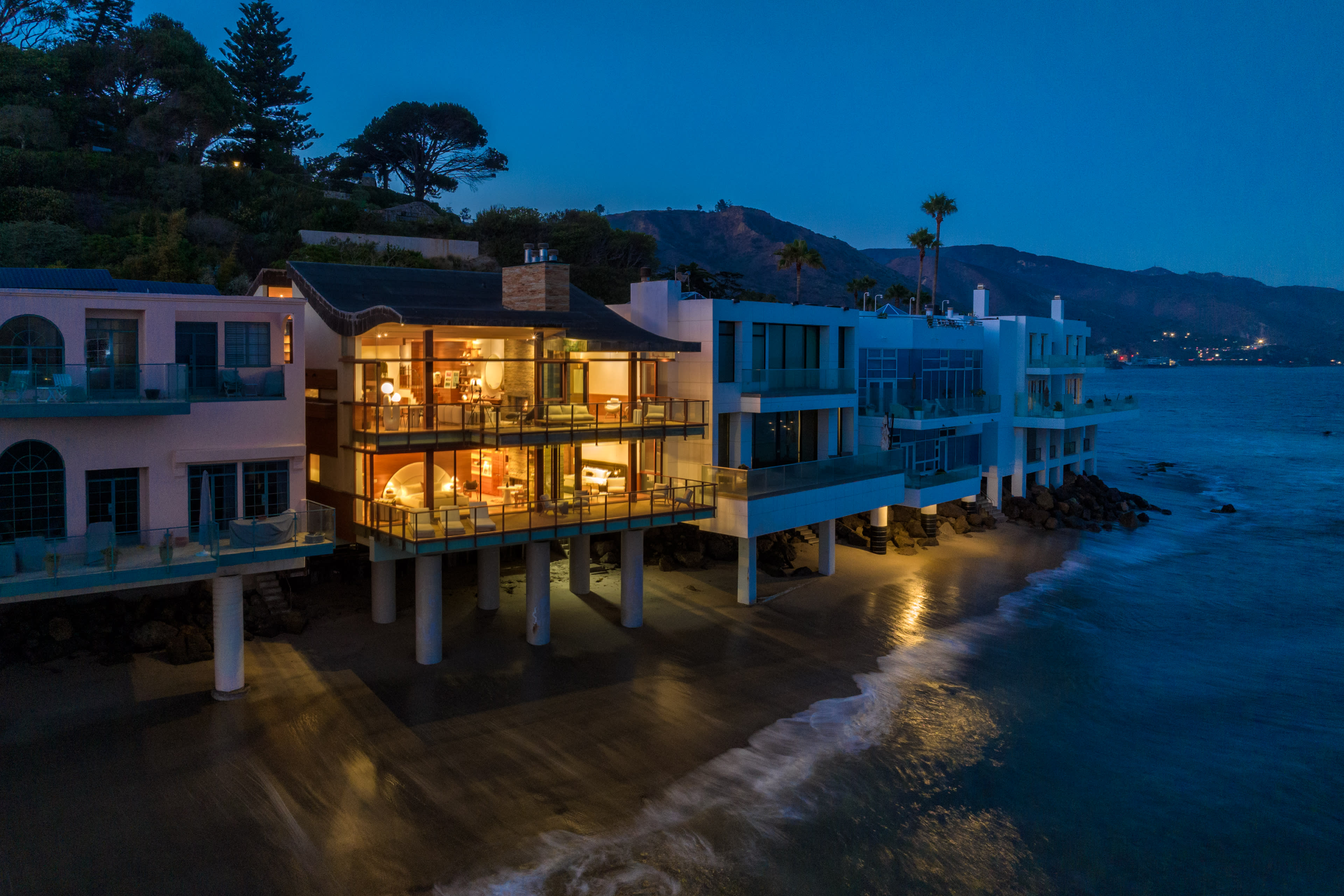 A modern beachfront house with large glass windows is illuminated at night, set against a mountainous backdrop by the sea.