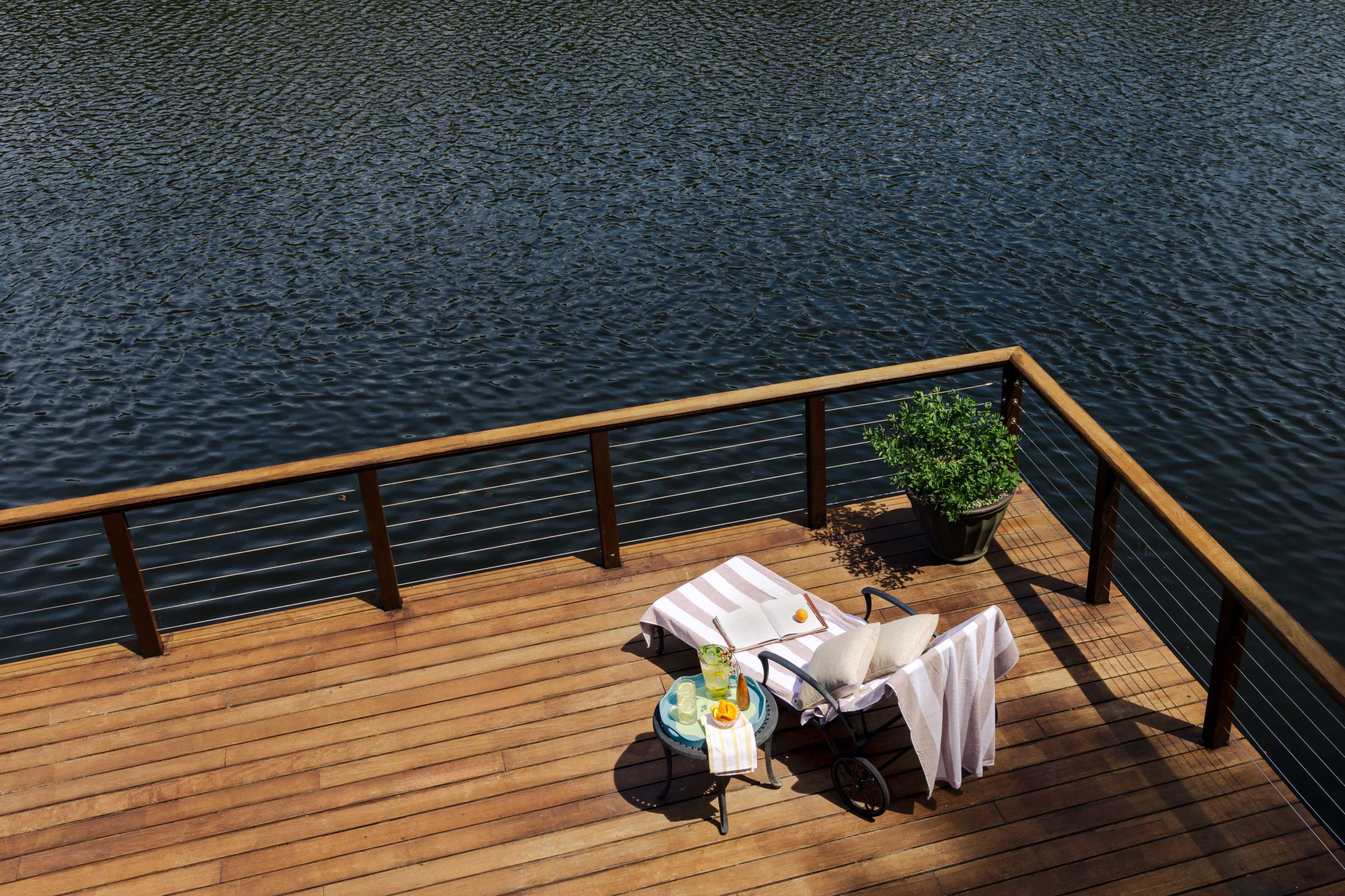 A wooden deck overlooking a body of water features a lounge chair and a small table with drinks and a plant.