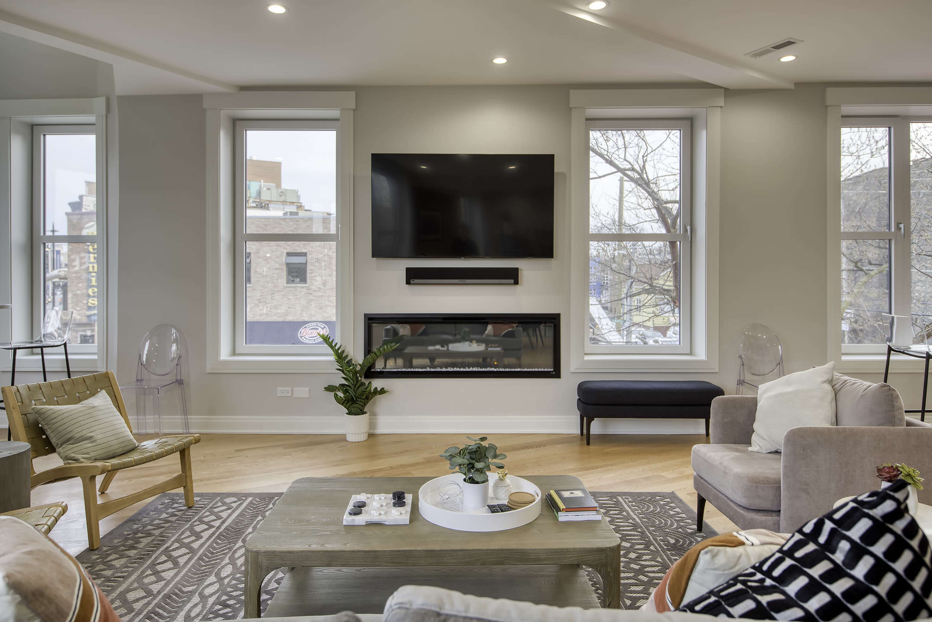 A modern living room with large windows, a wall-mounted television, and a contemporary fireplace surrounded by minimalist furniture.