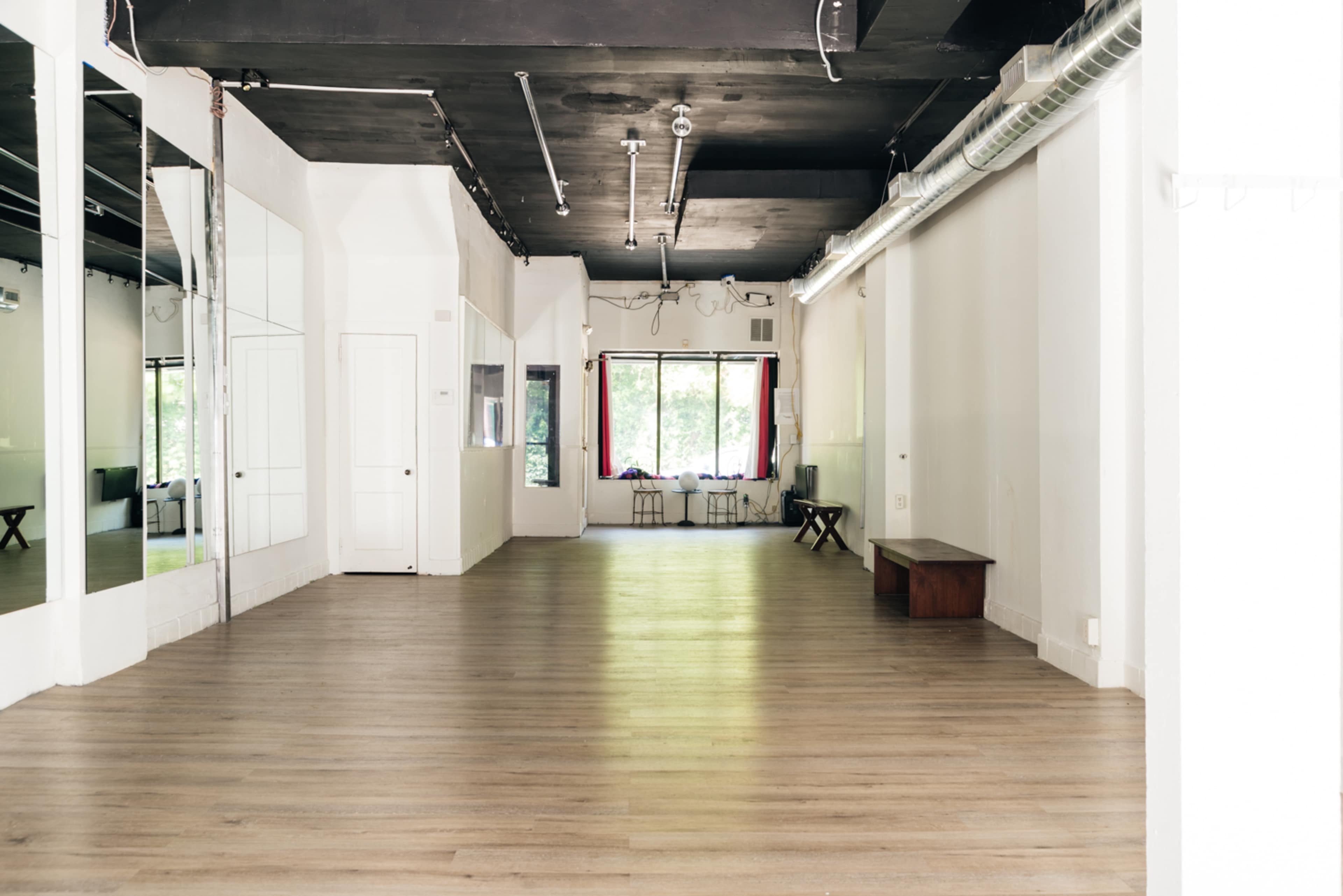 An empty dance studio with hardwood floors, large mirrors on one wall, and natural light streaming through a window at the far end.