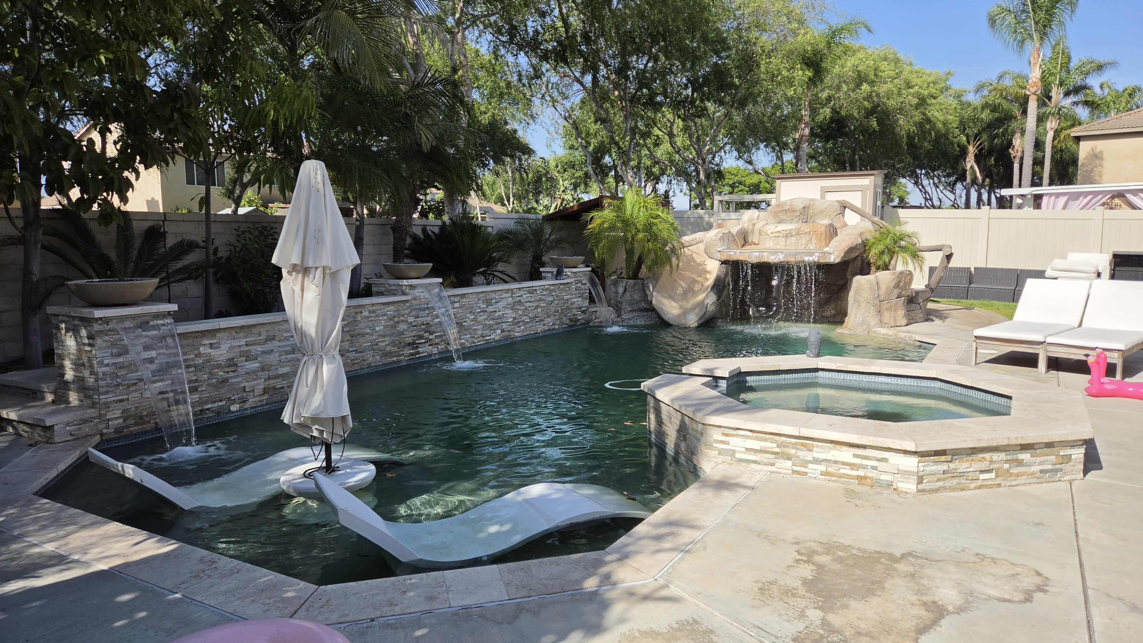 The image shows a backyard swimming pool with a waterfall feature, two lounge chairs, and an umbrella beside a hot tub.
