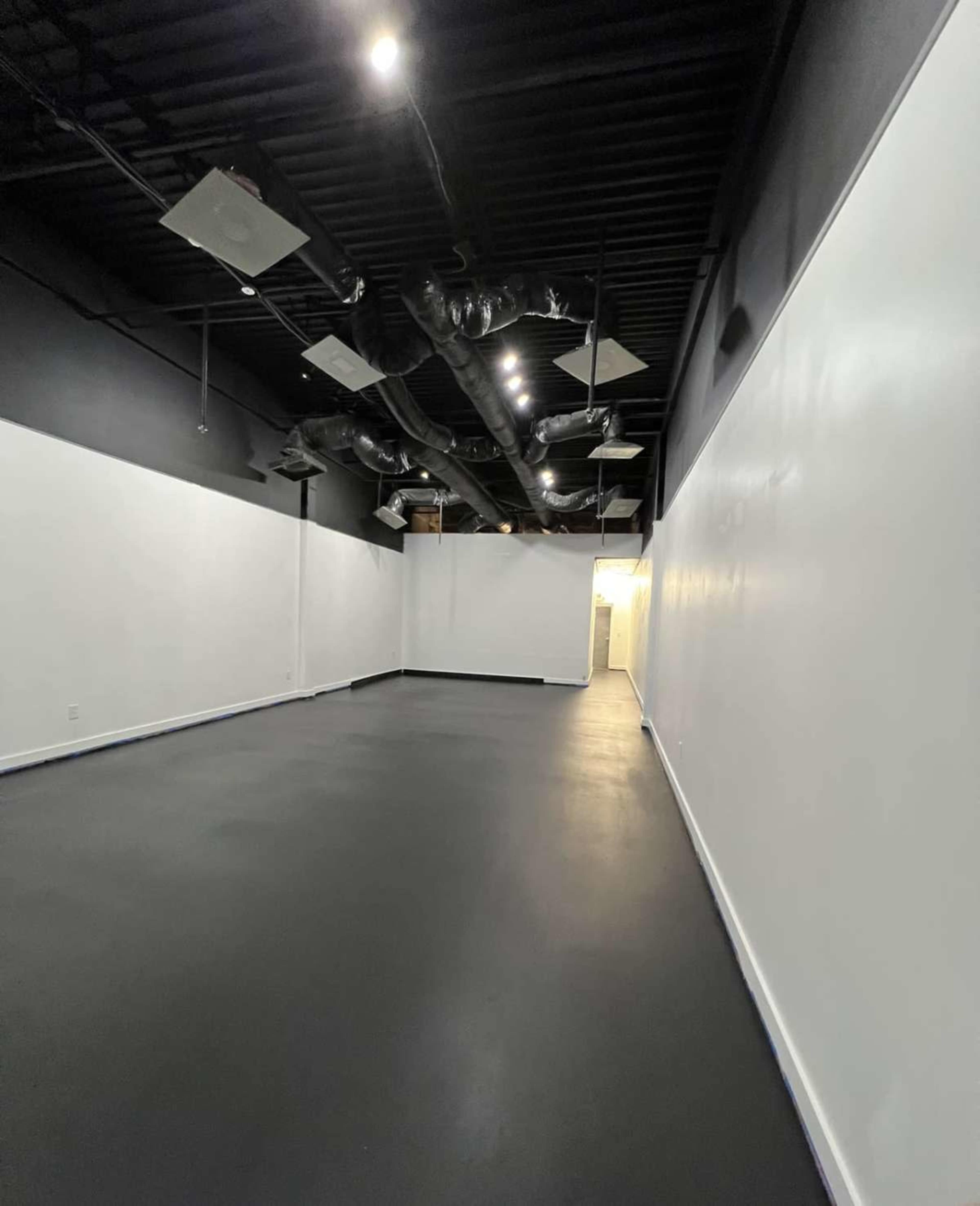 The image shows a spacious, minimally furnished room with black flooring and white walls, illuminated by ceiling lights and featuring exposed ductwork.